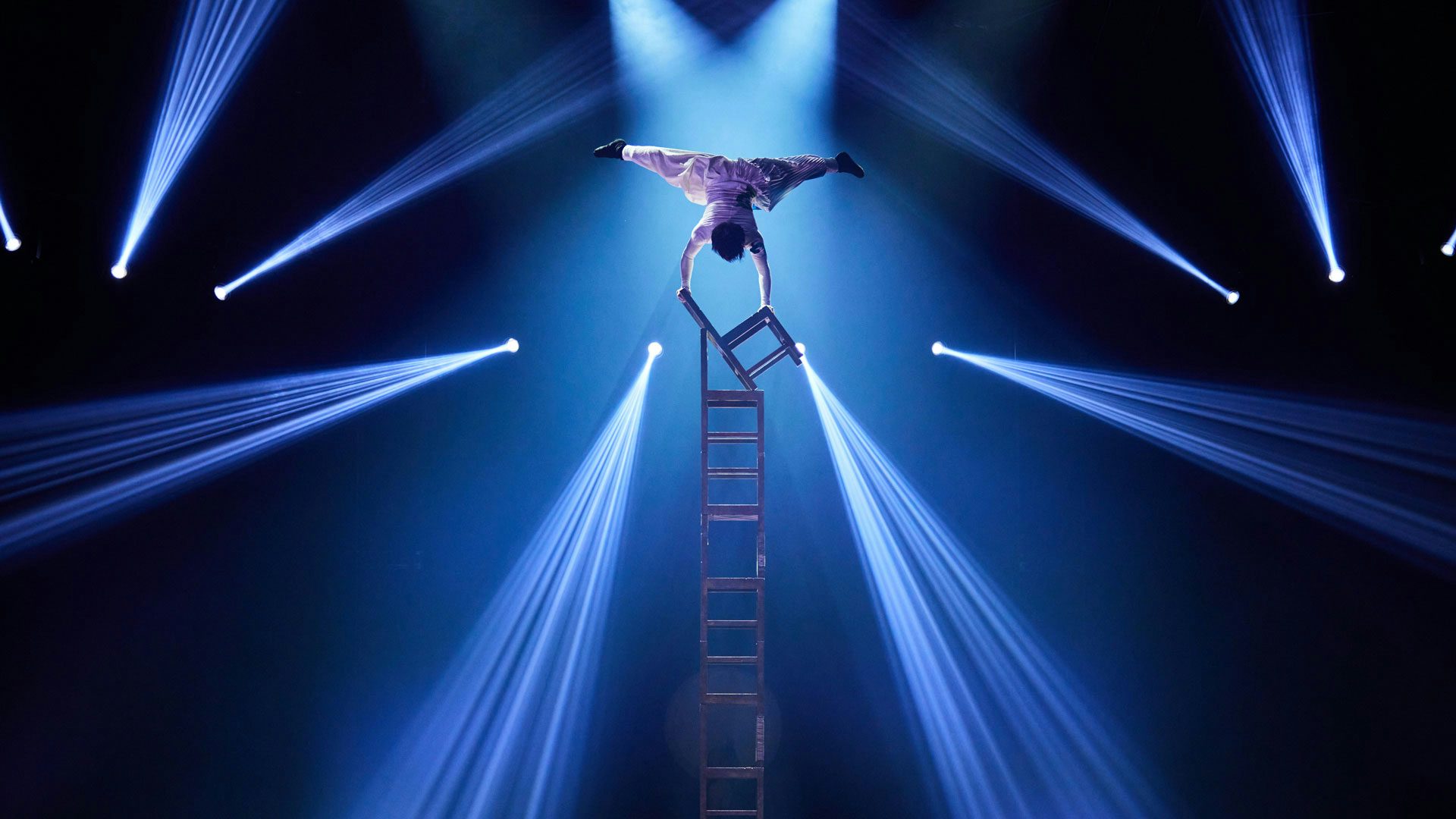 A performer in baggy white clothing with short dark hair does a handstand on a tower of six chairs under two spotlights on a stage. The performer holds both legs out in a straddle above their body. The background is dark and ten bright lights shine out in different directions.