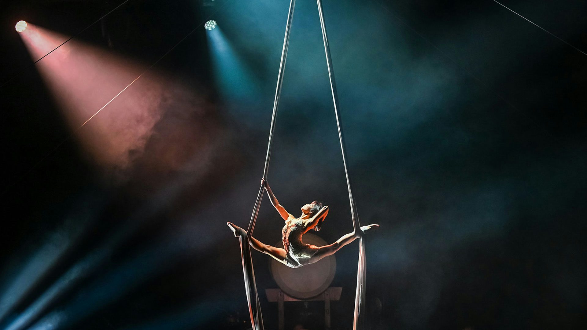 An aerialist hangs in a split between two silks wrapped around her ankles and suspended from the top of a dark, foggy stage. Two lights shine from the upper left corner of the stage.