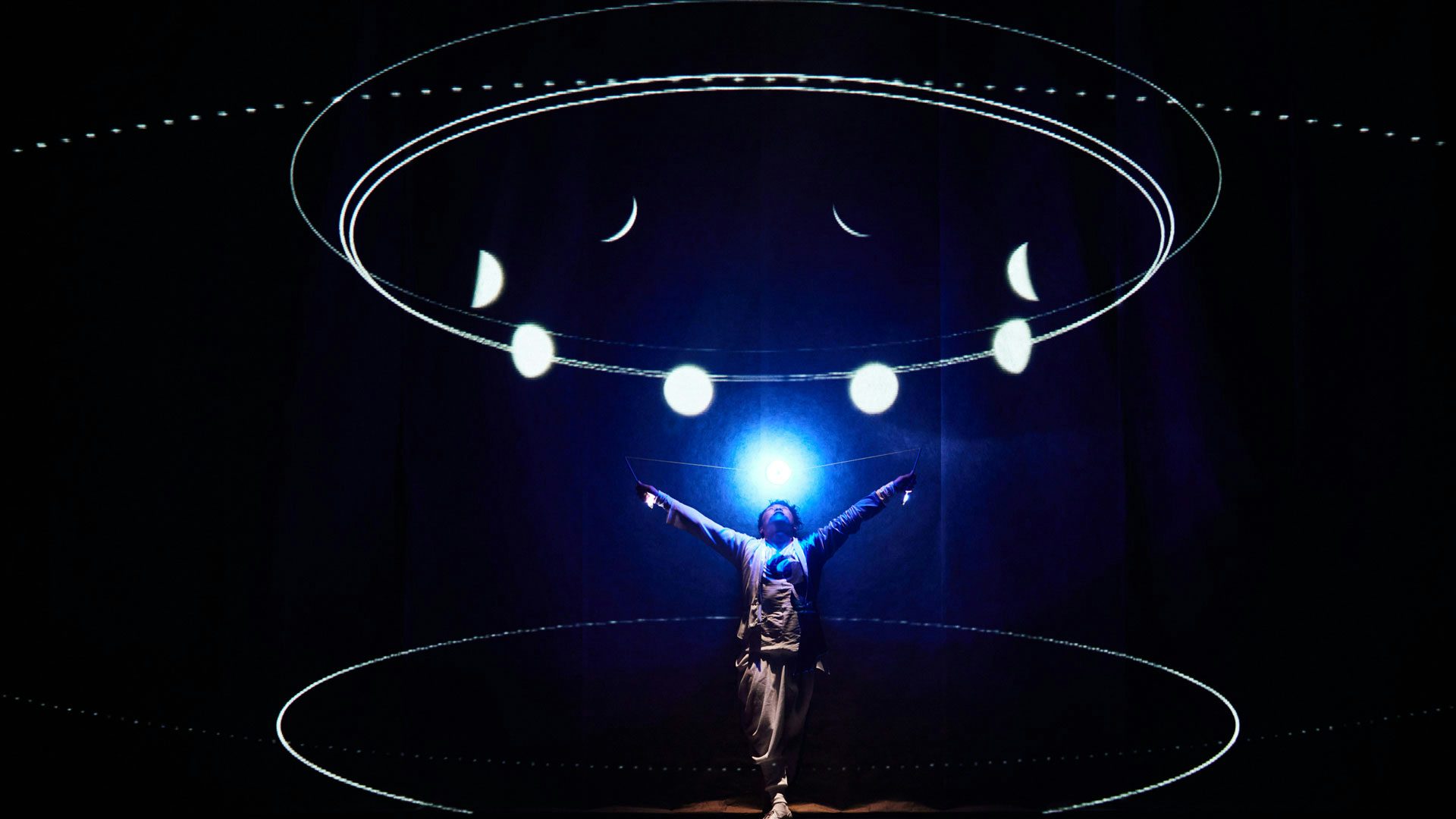 A performer with short black hair wears loosely fitting garments and balances a glowing diabolo above their head against a black background. White ellipses and the phases of the moon are projected on the performer and the dark stage.