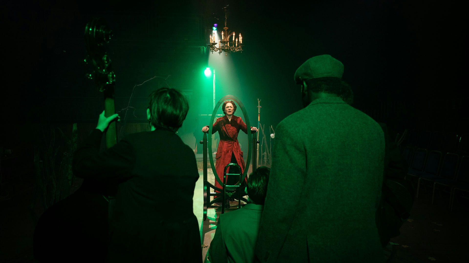 In the foreground, three performers wearing coats are covered in a green wash of dim light as they face away from the audience. In the center of a wooden stage, a spotlight shines on a woman with dark hair wearing a red, tufted velvet dressing gown. A gold chandelier hangs over her head. The woman peers straight ahead through the empty wooden frame of a full-length mirror as she grasps the frame on either side with both hands.