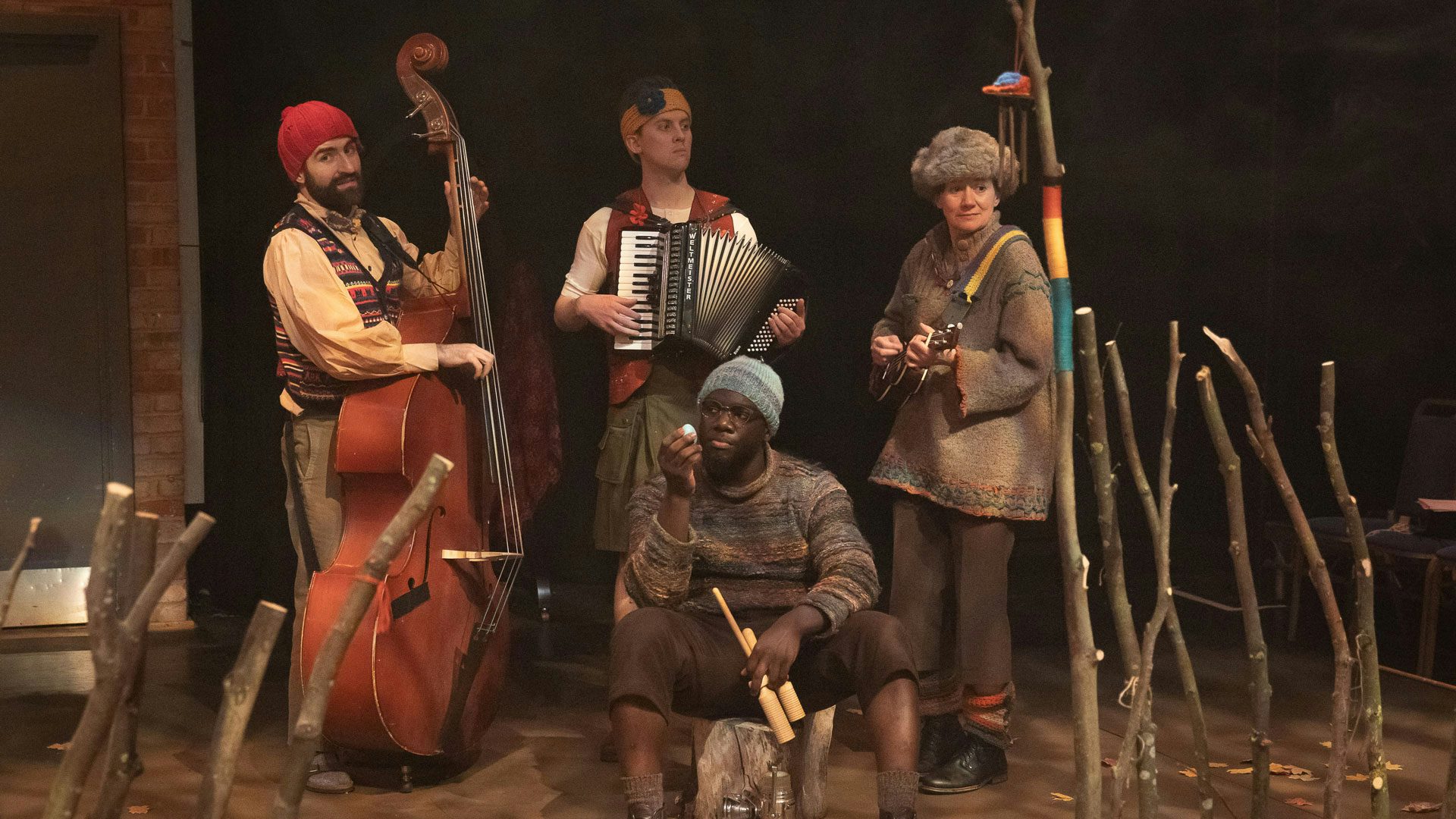 Ten large sticks are arranged like fences on either side of four performers. One performer wearing a blue knit cap, brown clothing and glasses sits on a block of wood and plays an egg shaker in one hand and holds a double guiro in the other. On the left, a performer with a dark beard wearing a red knit cap and a colorful sweater vest stands playing a double bass. In the center, a performer wearing earth-toned clothing and a yellow knit headband with a blue flower stands and plays a black and white accordion. On the right, a performer wearing a fur hat and loosely-fitting brown clothing plays a ukulele. The background is dark and hazy.