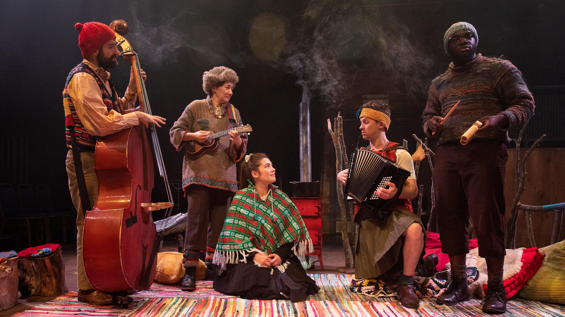 Three performers holding various instruments and dressed in earth tones and warm clothing stand around a young woman with dark hair sitting on the ground and wearing a green poncho adorned with tassels and a red and white pattern. Another performer with short brown hair and a yellow headband kneels beside the woman and plays a black accordion. The ground is covered by a multicolored rug and pillows.