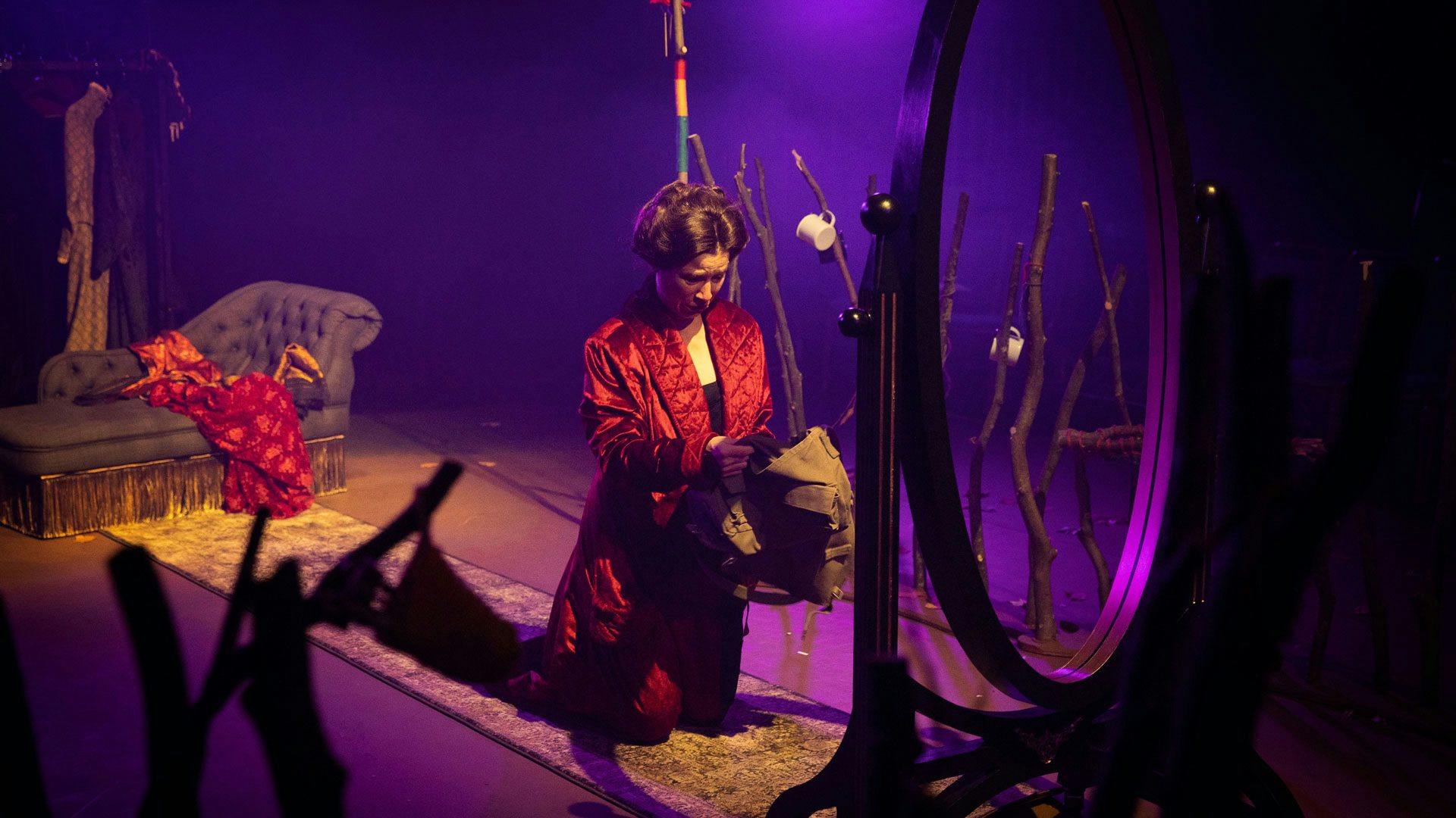 A woman with dark hair pulled back into an updo wearing a red, tufted, velvet dressing gown kneels before a wooden, full-length mirror frame and holds a satchel in soft yellow lighting. She peers into the satchel with a distressed look on her face. Fences of large sticks stand on either side of the woman. In the background drenched in purple lighting, a gray, upholstered chaise with gold fringe sits on a gray, patterned runner rug leading to the mirror frame.