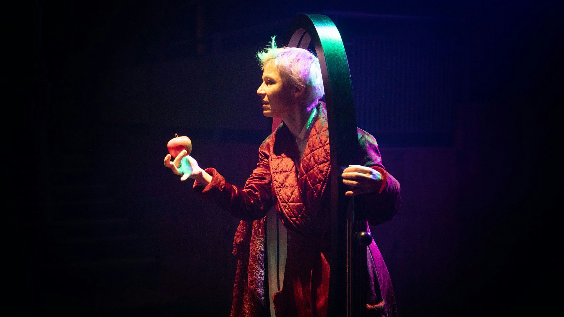 A woman with short white hair wearing a tufted, red velvet dressing gown holds a red apple in one hand as she peeks her head through the empty wooden frame of a standing full-length mirror against a black background. She holds the edge of the wooden mirror frame with the other hand.