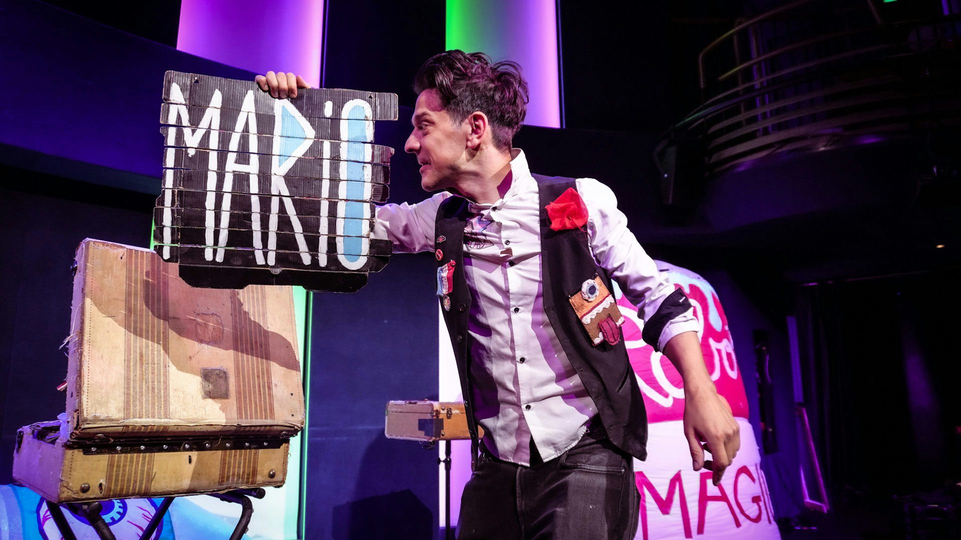 A man with dark hair wearing a gray button-down shirt, black pants and a black vest adorned with buttons, a striped medal and a fabric face with teeth, a tongue and one blue eyeball stands on a stage and holds a black cardboard sign to the left of his face. He looks at the sign, which has "Mario" painted in white letters and light blue accents. The man stands in front of two beige trunks.