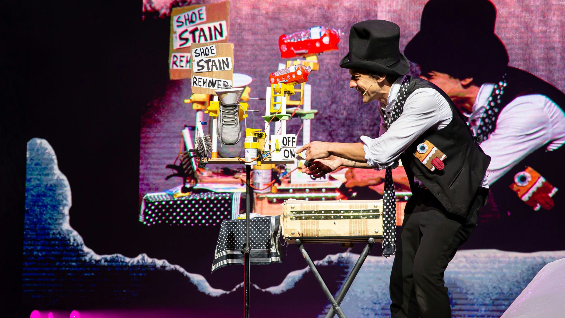 A man wearing a black top hat, a gray shirt, black pants, a black tie with white polka dots and a black vest adorned with a fabric face with teeth, a tongue and one blue eyeball stands on a stage beside an apparatus made of household objects. The apparatus has a cardboard sign that reads "Shoe Stain Remover," a gray sneaker suspended in the air with a white funnel in it and a bristled cleaning brush. On the back right corner of the apparatus stand, a plastic bottle of water is suspended in the air by a red dish, and below it, there’s a white on/off switch with black handwritten letters. The man points to the "on" switch. In the background, a large screen displays a live video feed of the stage.