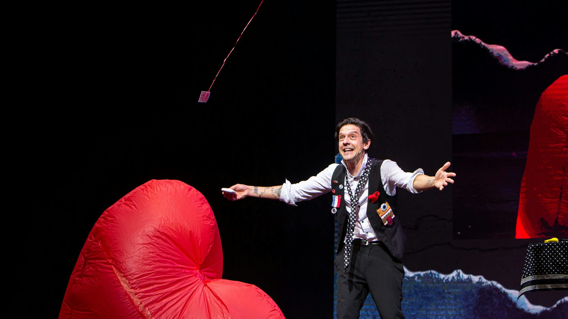 A man with dark hair wearing a gray button-down shirt, black pants, a black tie with white polka dots and a black vest adorned with buttons, a striped medal and a fabric face with teeth, a tongue and one blue eyeball stands on a stage with his arms outstretched to either side, holding a deck of playing cards in one hand. An inflatable red organic shape sits in the bottom left corner. A red string enters from the ceiling and holds a red playing card in midair.