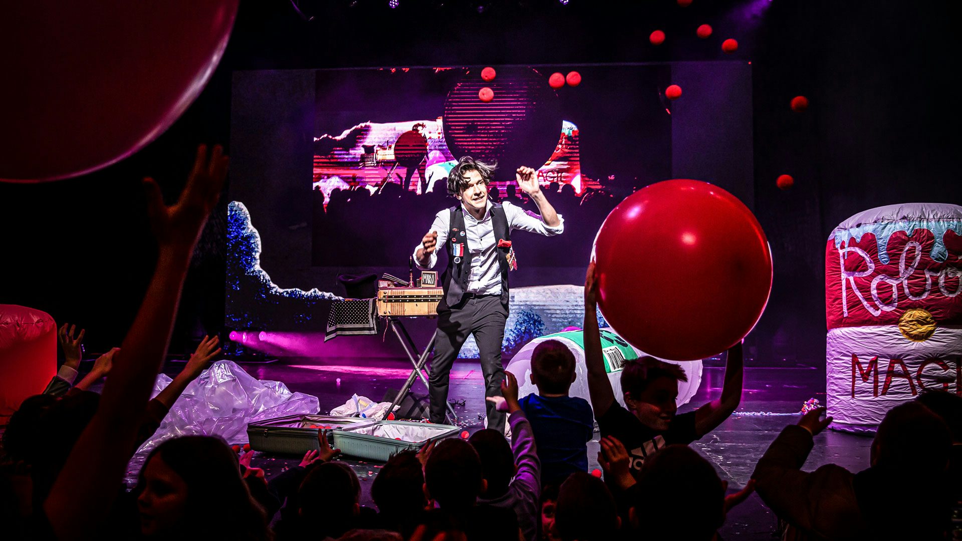 A man with dark hair wearing a gray button-down shirt, black pants and a black vest adorned with buttons, a striped medal and a face with teeth and a red tongue stands on a stage with his hands raised near his face behind a large unzipped suitcase on the ground. The open suitcase is partially filled with small red balls. Several small red balls fall from the ceiling and the audience of children in the foreground raise their arms in the air to catch two large red balls. In the background, there is a large screen with a live video feed of the stage.
