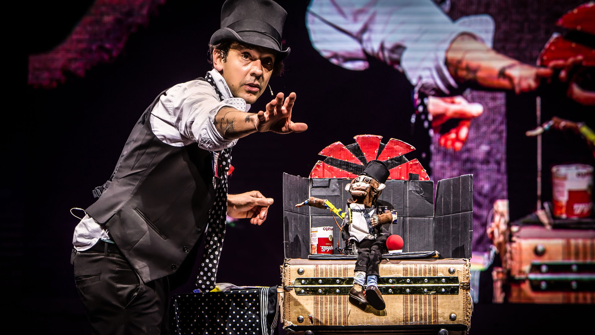 A man wearing a black top hat, vest, gray shirt, black pants, and black and white polka dotted tie faces the right and holds one hand straight out to the audience. In front of him, a robot monkey wearing the same outfit sits on a trunk in front of a black and red cardboard backdrop and mimics the man's arm position. Behind them both, a large screen shows a live video feed of the stage.