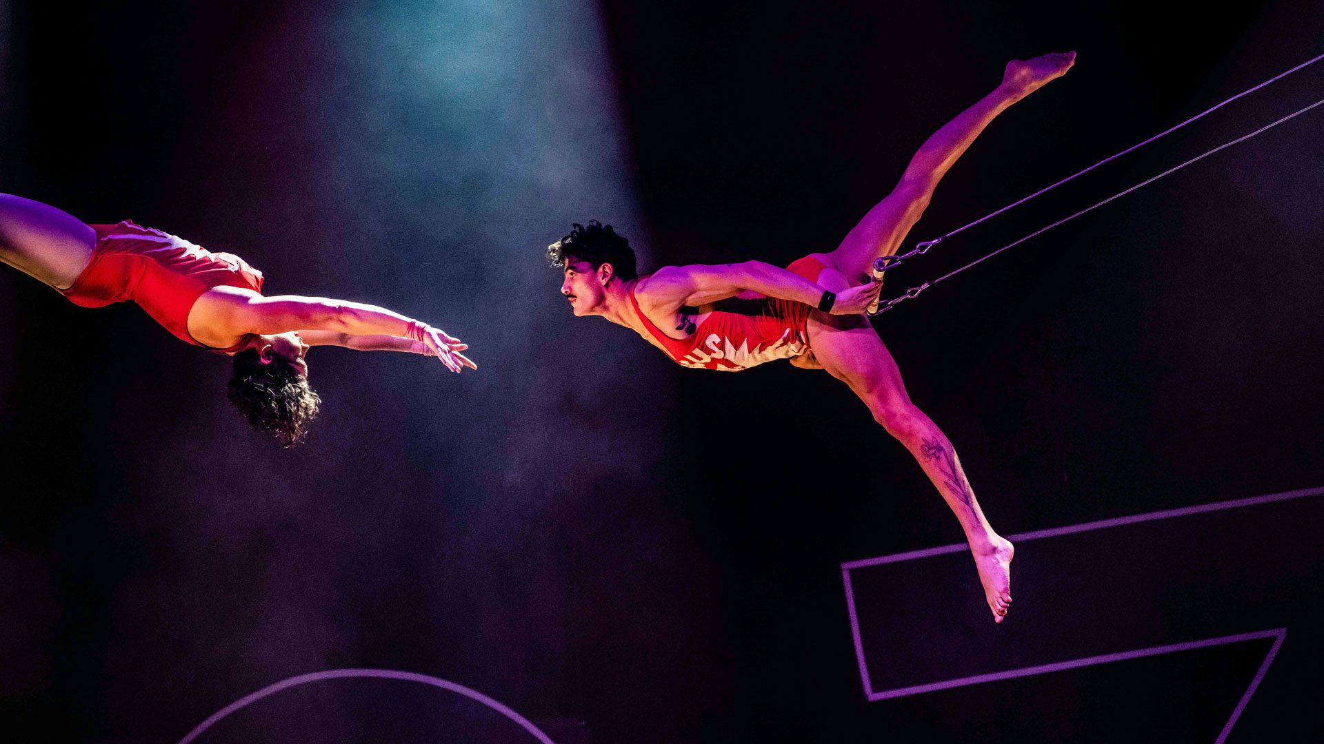 Against a foggy black background, two trapeze performers with short dark curly hair wearing red unitards swing towards one another in the air. The performer on the left faces up with their arms outstretched towards the performer on the right. The performer on the right holds onto the trapeze bar with their hand and stretches their legs in a v shape behind them.
