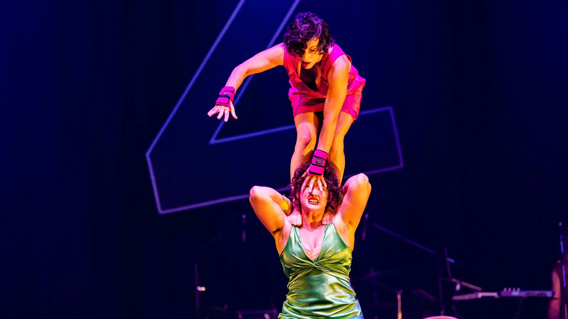 A woman with dark curly hair wearing a short, bright pink dress and wrist guards bends down and stands on the shoulders of a woman with dark curly hair wearing a bright green silky dress against a dark background. The woman in the green dress makes a pained face, which is partially covered by the woman in the pink dress’s hand.