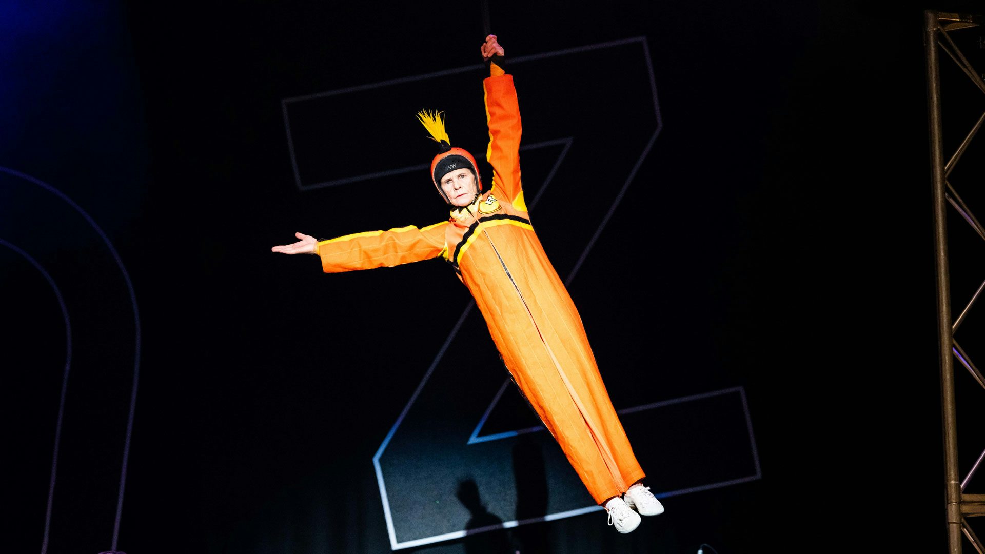 A performer wearing an orange coverall suit with black and yellow stripes, an orange helmet with a yellow mohawk and white sneakers hangs midair from a black piece of fabric with one hand. A spotlight shines on her as she reaches out with her other hand and holds her legs together straight below her against a black background.