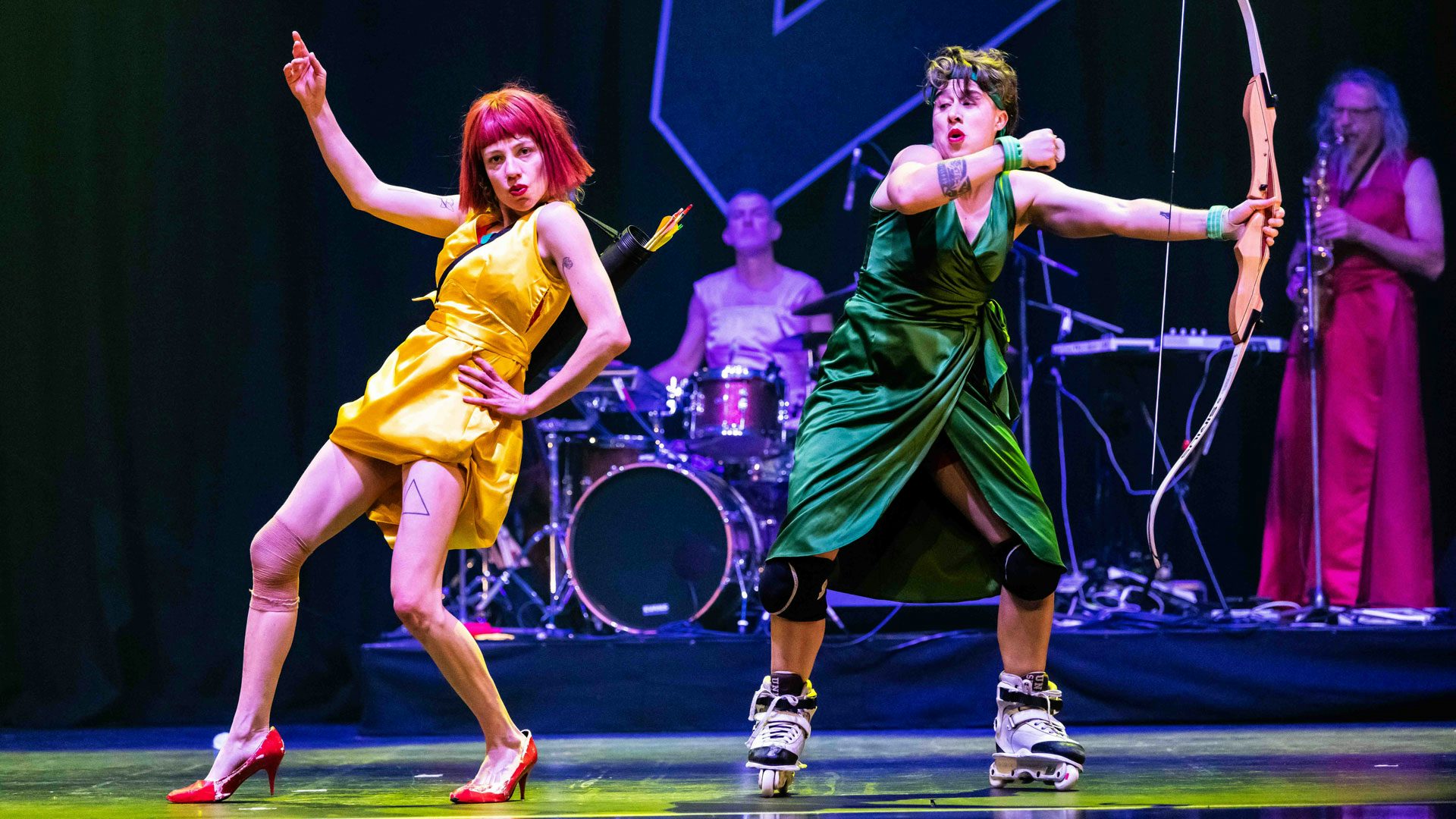 Two women wear red lipstick and dance on stage. The woman on the left has a short red bob and wears a short yellow dress, an archery quiver and red high heels. The woman on the right has dark hair and wears an emerald green dress, green bracelets, a green headband and roller blades. The woman on the right holds an archer's bow in one hand. In the background, a drummer and saxophonist wear pink dresses.