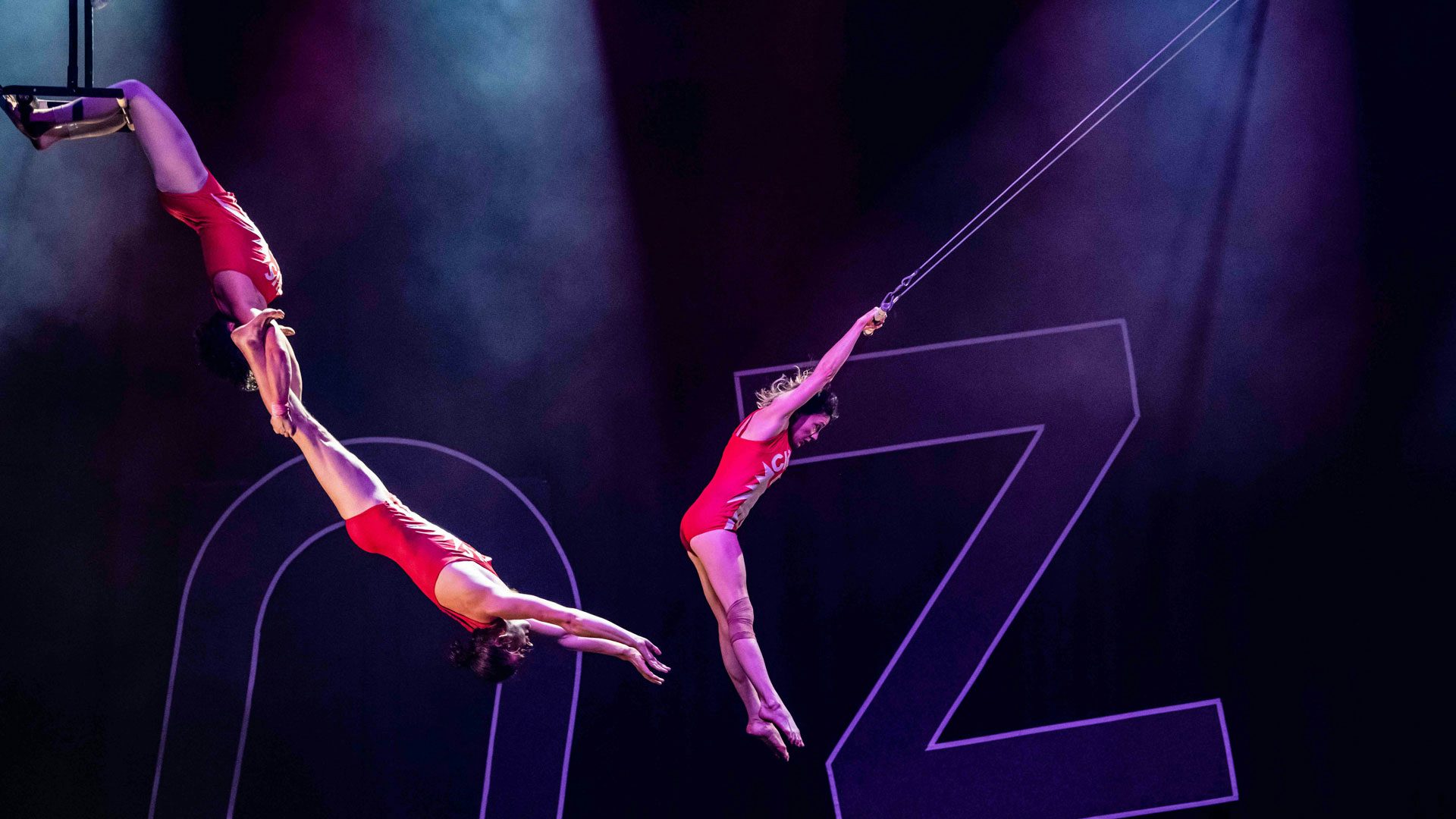 Two women wear red lipstick and dance on stage. The woman on the left has a short red bob and wears a short yellow dress, an archery quiver and red high heels. The woman on the right has dark hair and wears an emerald green dress, green bracelets, a green headband and roller blades. The woman on the right holds an archer's bow in one hand. In the background, a drummer and saxophonist wear pink dresses.