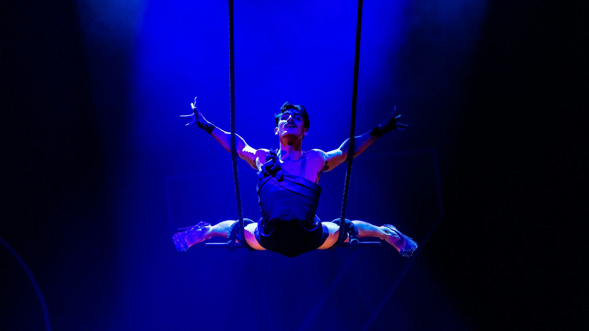 A man with short dark hair and a thin dark mustache stretches his arms and legs out behind him on a trapeze bar. The lighting is a deep blue. The man wears sparkly high-heeled shoes and a one-shoulder mini dress.