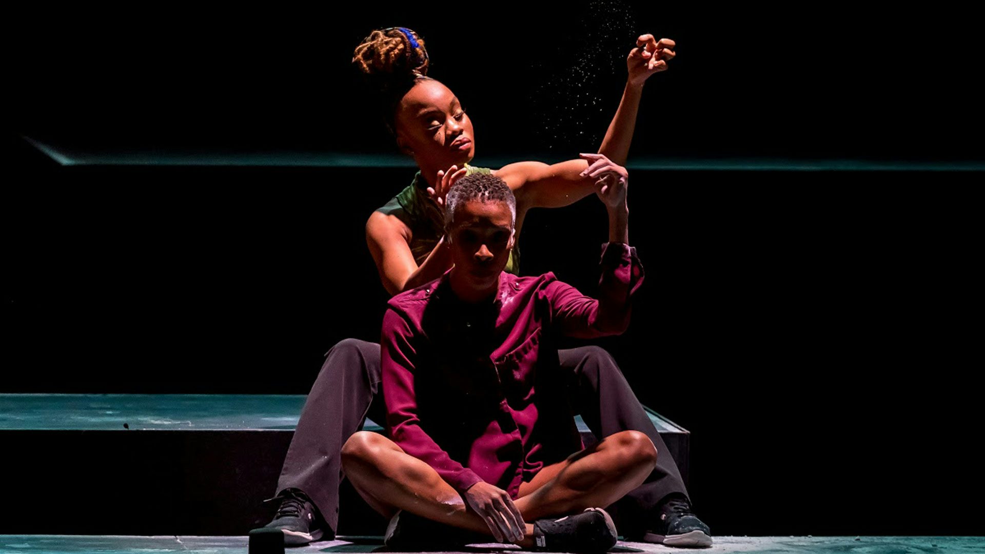 Two Black women sit on a dimly lit stage against a black background. One woman has short hair and wears a maroon shirt with long sleeves. She sits in front of the other woman with her legs crossed and raises one arm above her head. The second woman wears a green tank top and wears her hair in a top knot. She sits on a raised part of the stage with her legs spread to either side and holds one hand behind the other woman's head and one arm raised above her head.