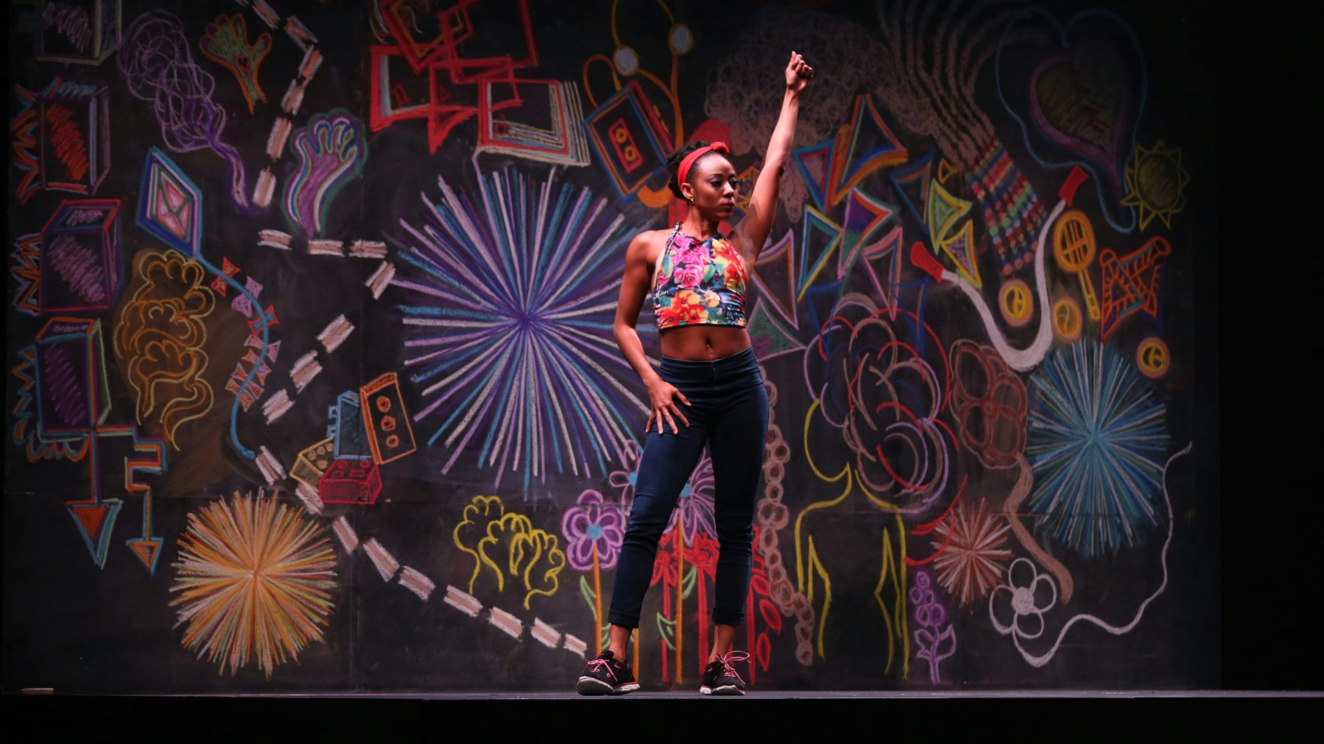 A thin Black woman in a floral tank top, red headband, black sneakers and mid-rise skinny jeans stands on a stage with her feet apart, one hand on her hip and one hand raised above her head in a fist. The background is covered in colorful chalk drawings.
