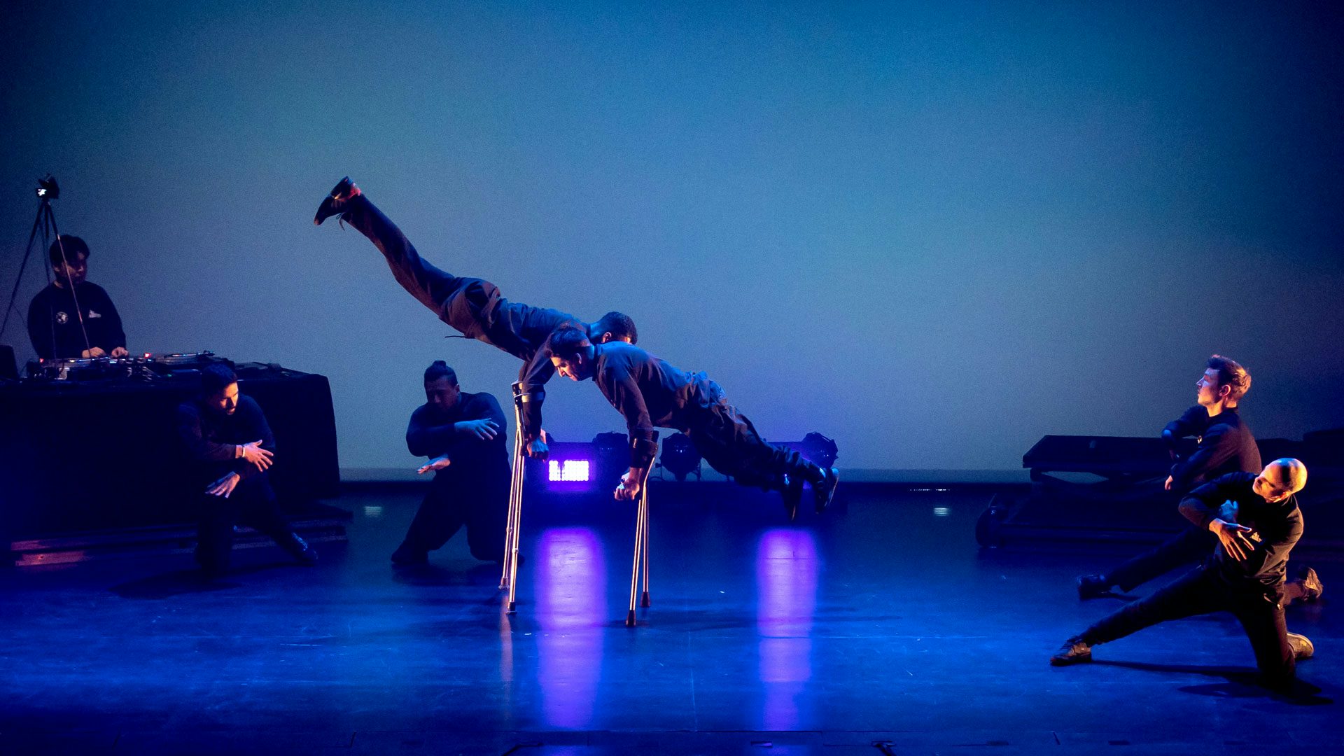 On a dark stage with blue lighting, two men dressed in black balance in handstands on forearm crutches. Four men dressed in black kneel in a circle around them with their arms crossed. In the background to the left, a DJ stands behind a turntable.