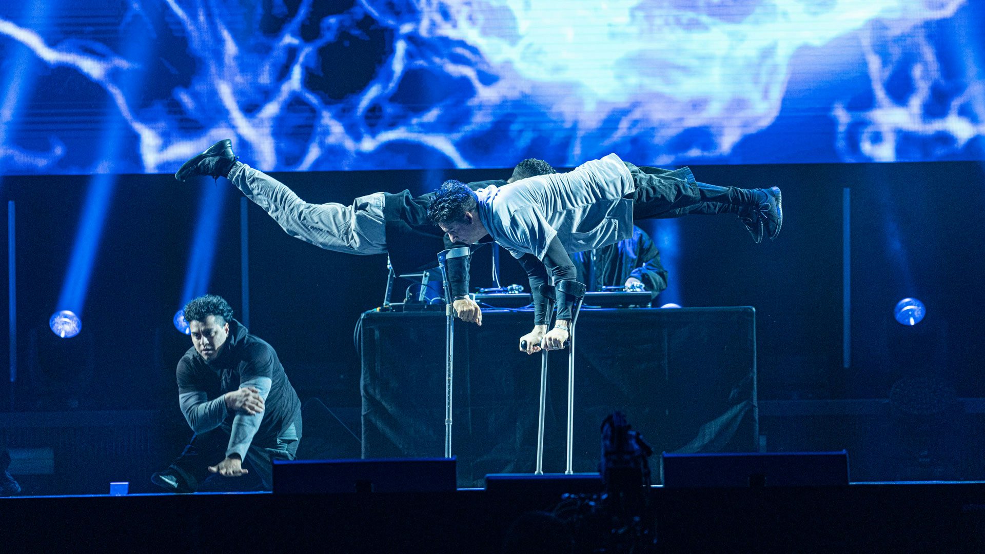 On a dark stage with a blue, white and purple lightning backdrop, two men dressed in black and gray balance in handstands on forearm crutches. Behind them, a man with dark curly hair kneels down. In the background, a DJ stands behind a turntable.