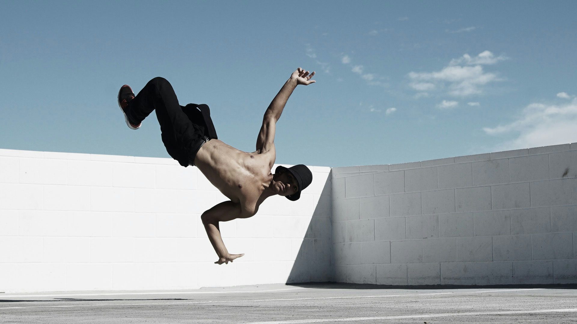 On a parking garage roof, a man wearing a black bucket hat, pants and sneakers strikes a pose in midair with his feet and one arm outstretched to the sky and one arm bent at a right angle, palm flat and facing the ground. The background shows a white cement brick wall and a cloudy, pale blue sky.