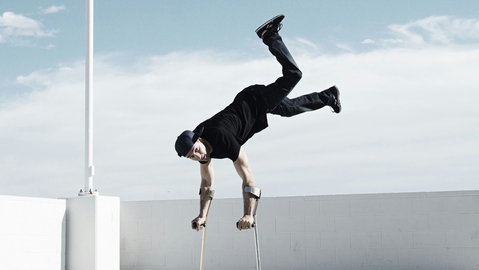 On a parking garage roof, a man wearing a black sideways cap, t-shirt, sneakers and jeans balances in a handstand on two forearm crutches. The background shows a white cement block wall and a cloudy pale blue sky.