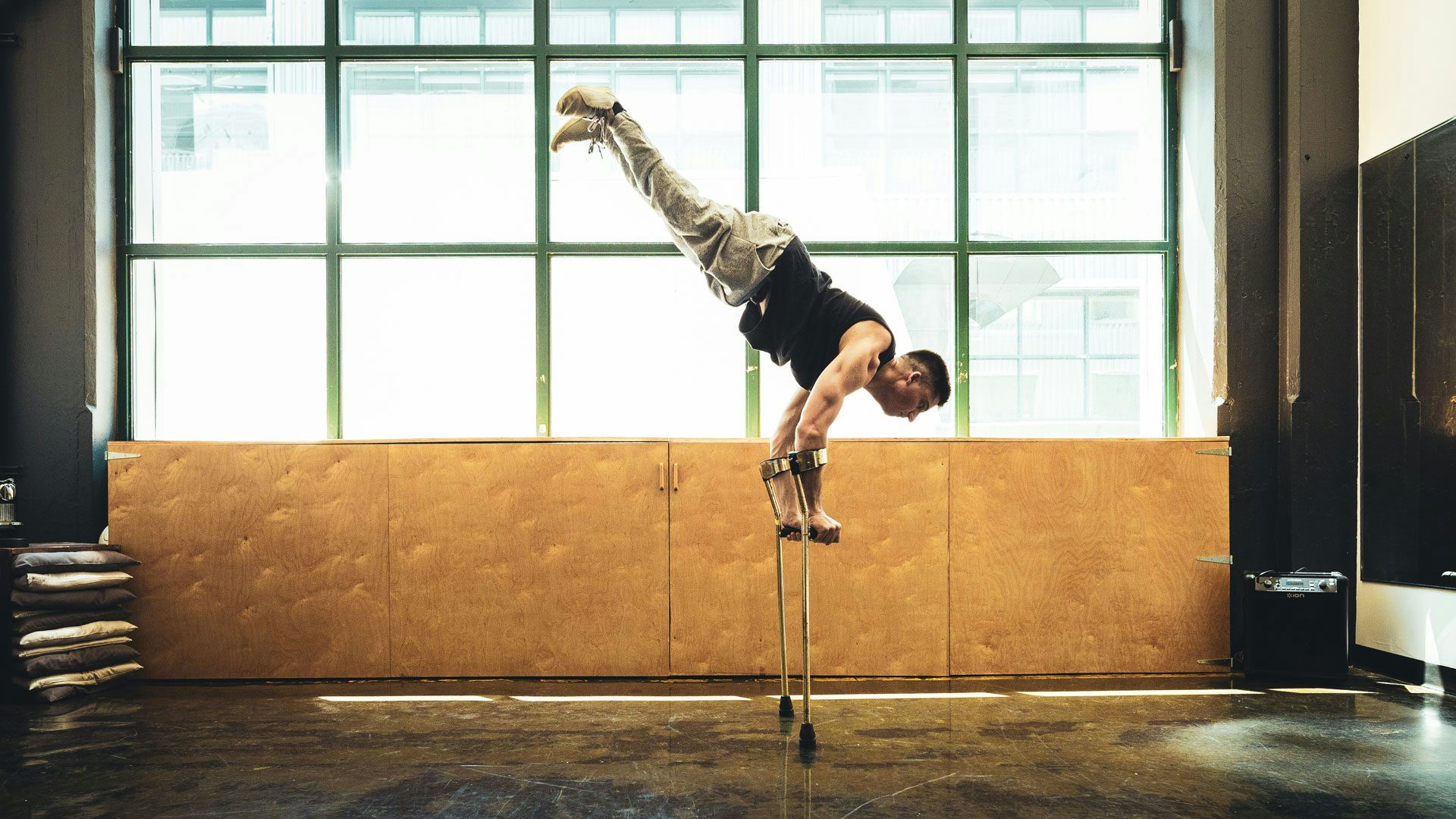 In an empty room with big warehouse windows, a man with short dark hair wearing a black tank top, gray sweatpants and white sneakers balances in a handstand on two forearm crutches.