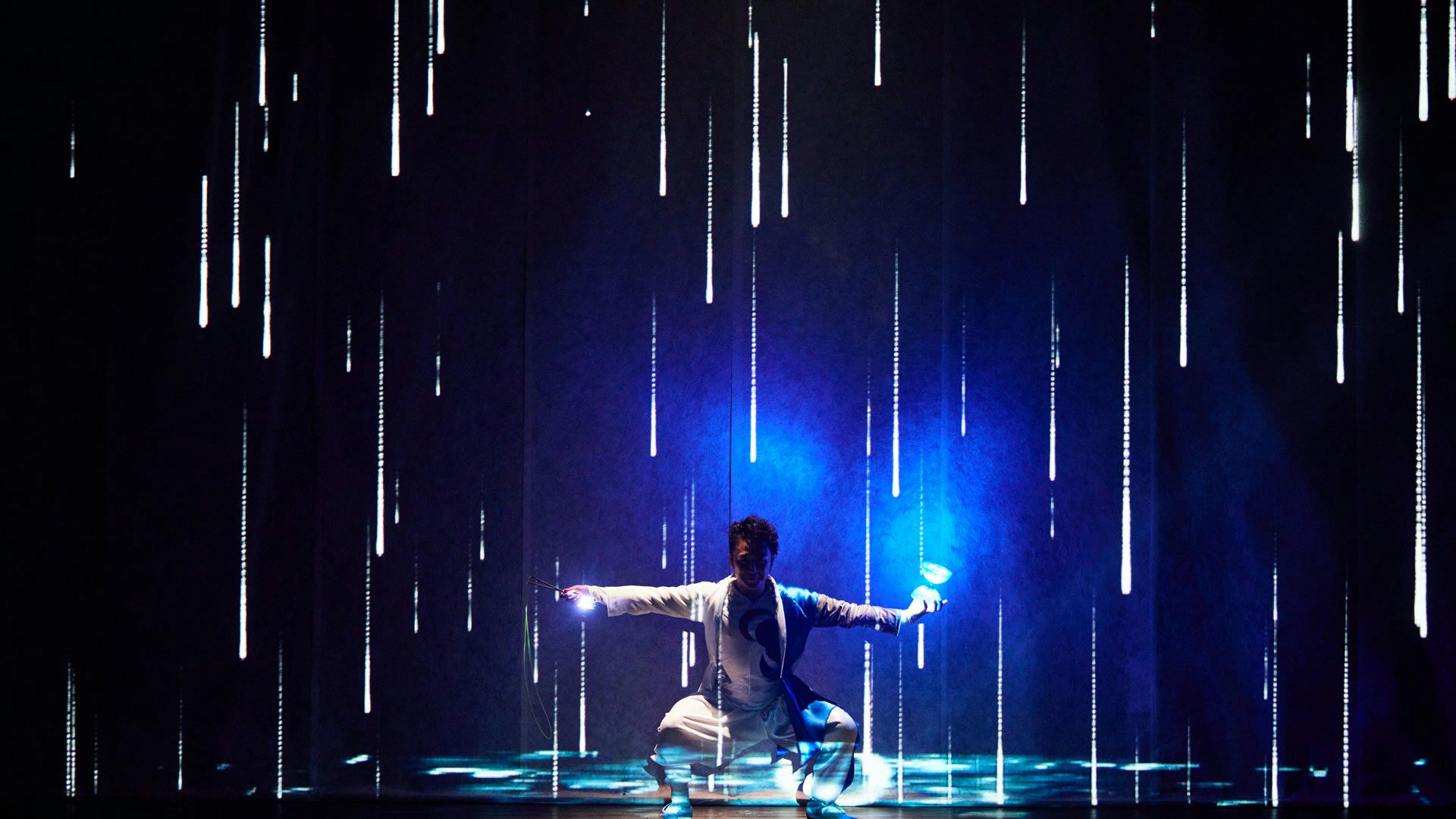 A performer with short black hair in a loose-fitting white outfit squats with their arms and legs out to either side of their body. The performer holds a glowing diabolo in one hand and a glowing diabolo string in the other hand. Falling rain and puddles are projected on the performer and the dark background on the stage.