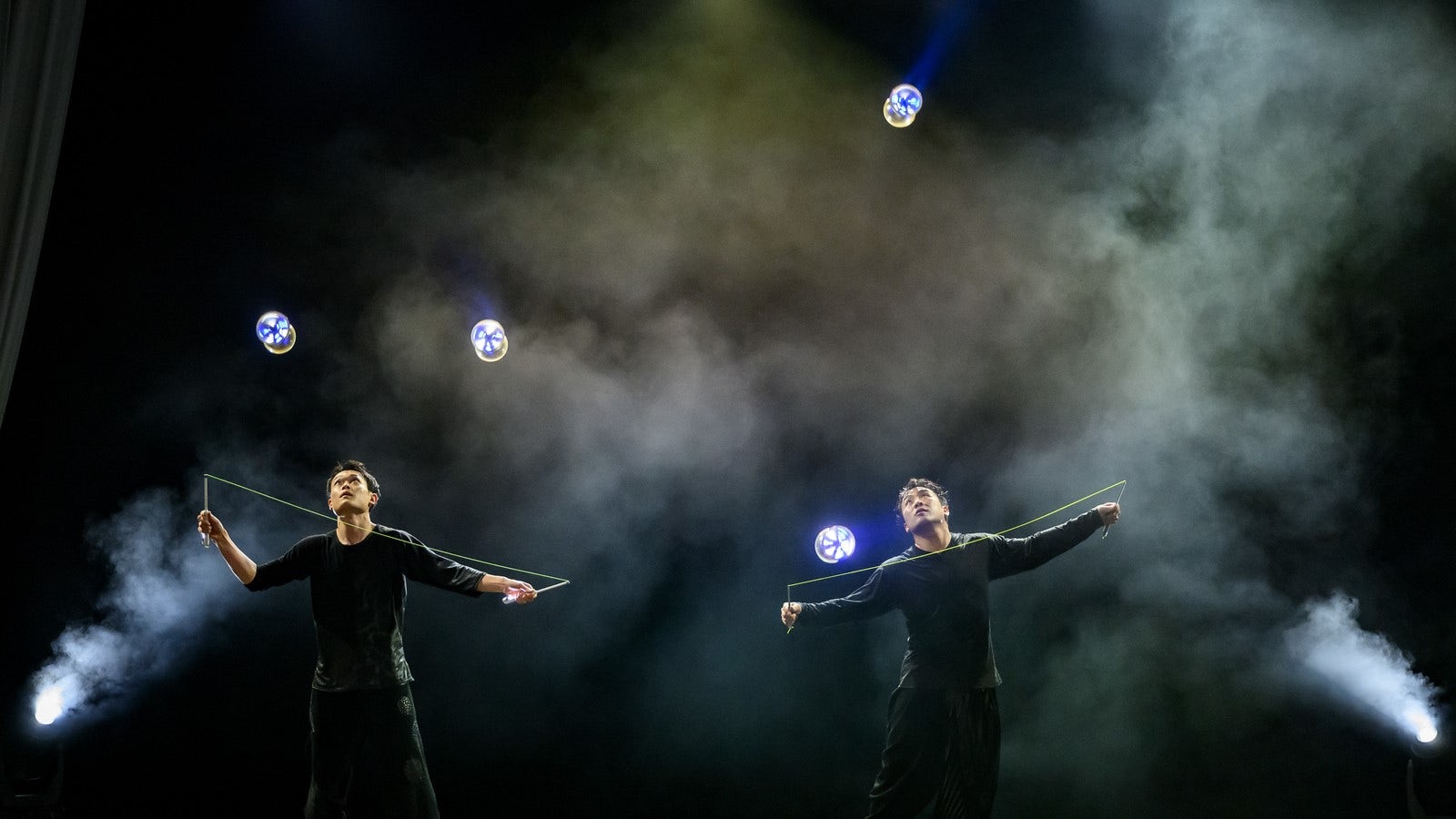 Two performers wearing black clothes use two glowing blue diablos against a black and smoky background.