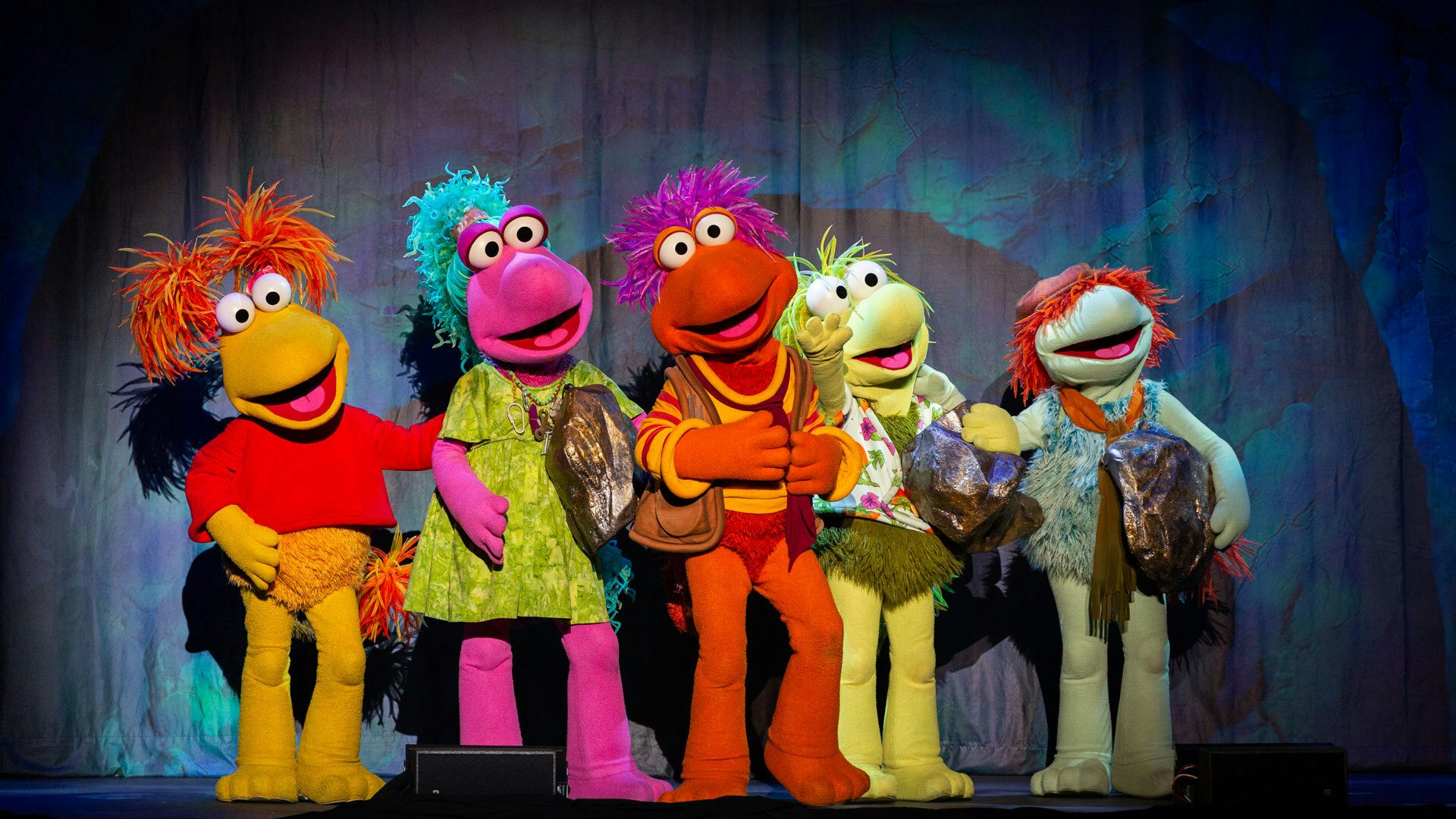 On a stage with a cave backdrop, five colorful and furry Fraggles stand in a line with their mouths open. From left to right, the Fraggles are yellow, pink, orange, yellow-green and teal. The pink, teal and yellow-green Fraggles hold large rocks.