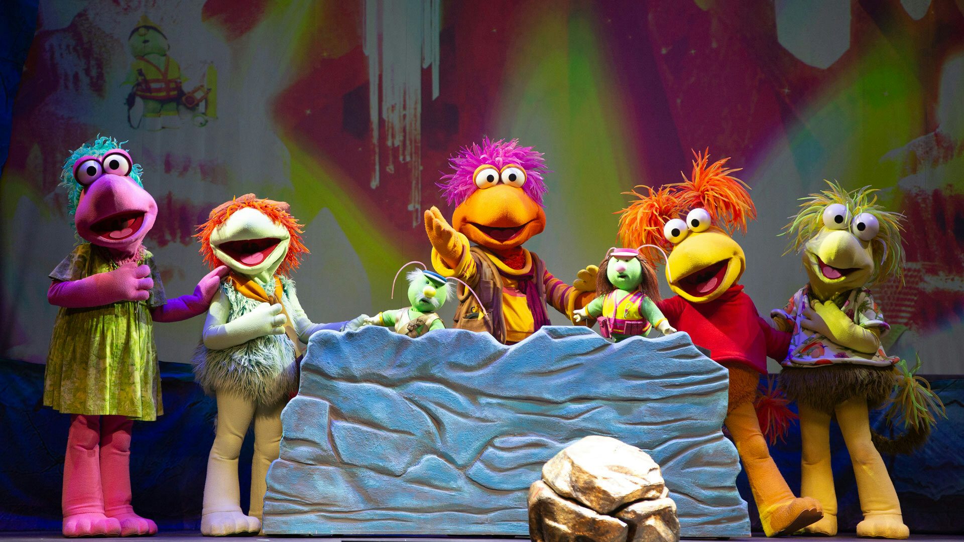 On a stage with a red and yellow background and rock set pieces, five colorful Fraggles stand with their mouths open near two small green puppets with antennae. The pink, teal and yellow-green Fraggles hold one hand over their chest. The yellow Fraggle holds its arms out, and the orange Fraggle in the middle holds their arm out to the audience.