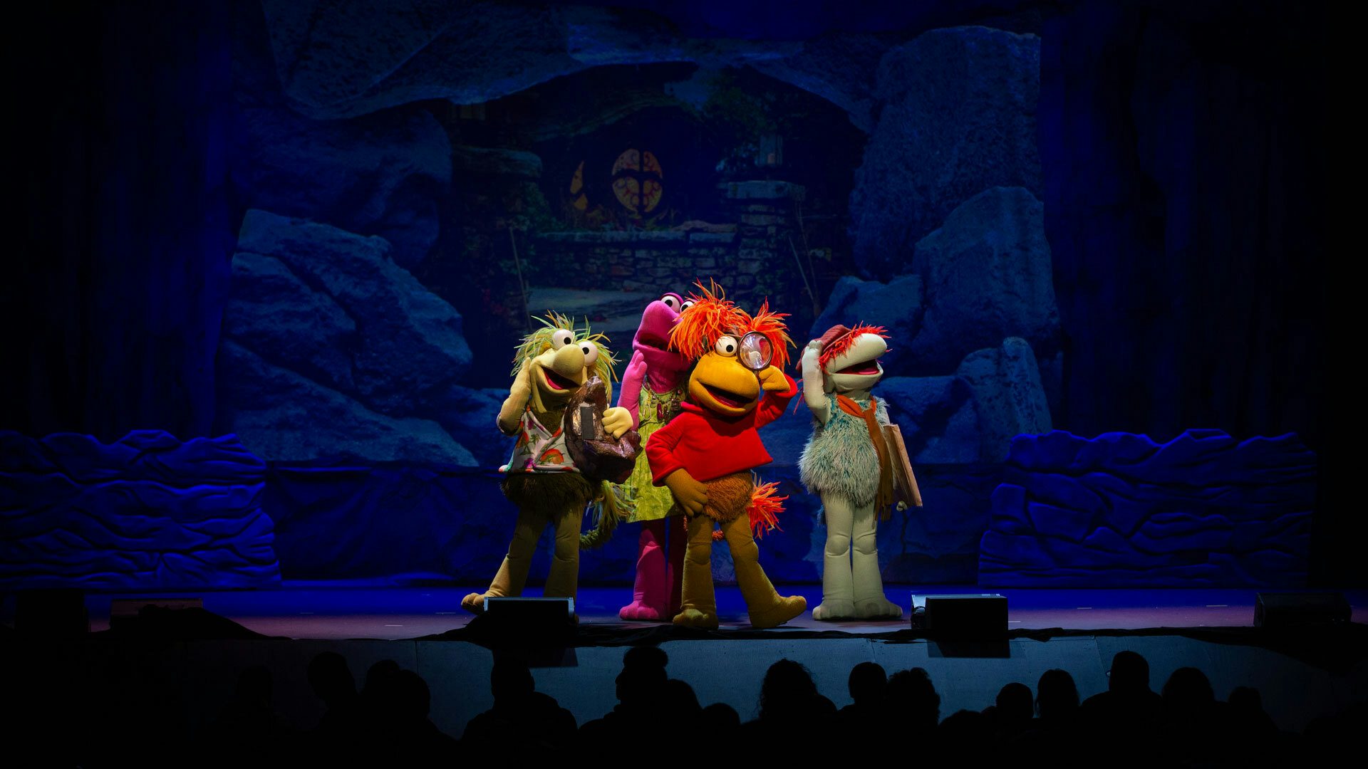 On a stage with a blue cave backdrop, four colorful, fuzzy Fraggles stand in a group facing the audience. The yellow Fraggle in front holds a magnifying glass. The yellow-green, pink and teal Fraggles surrounding the yellow one hold their hands up to their faces and head and look around.