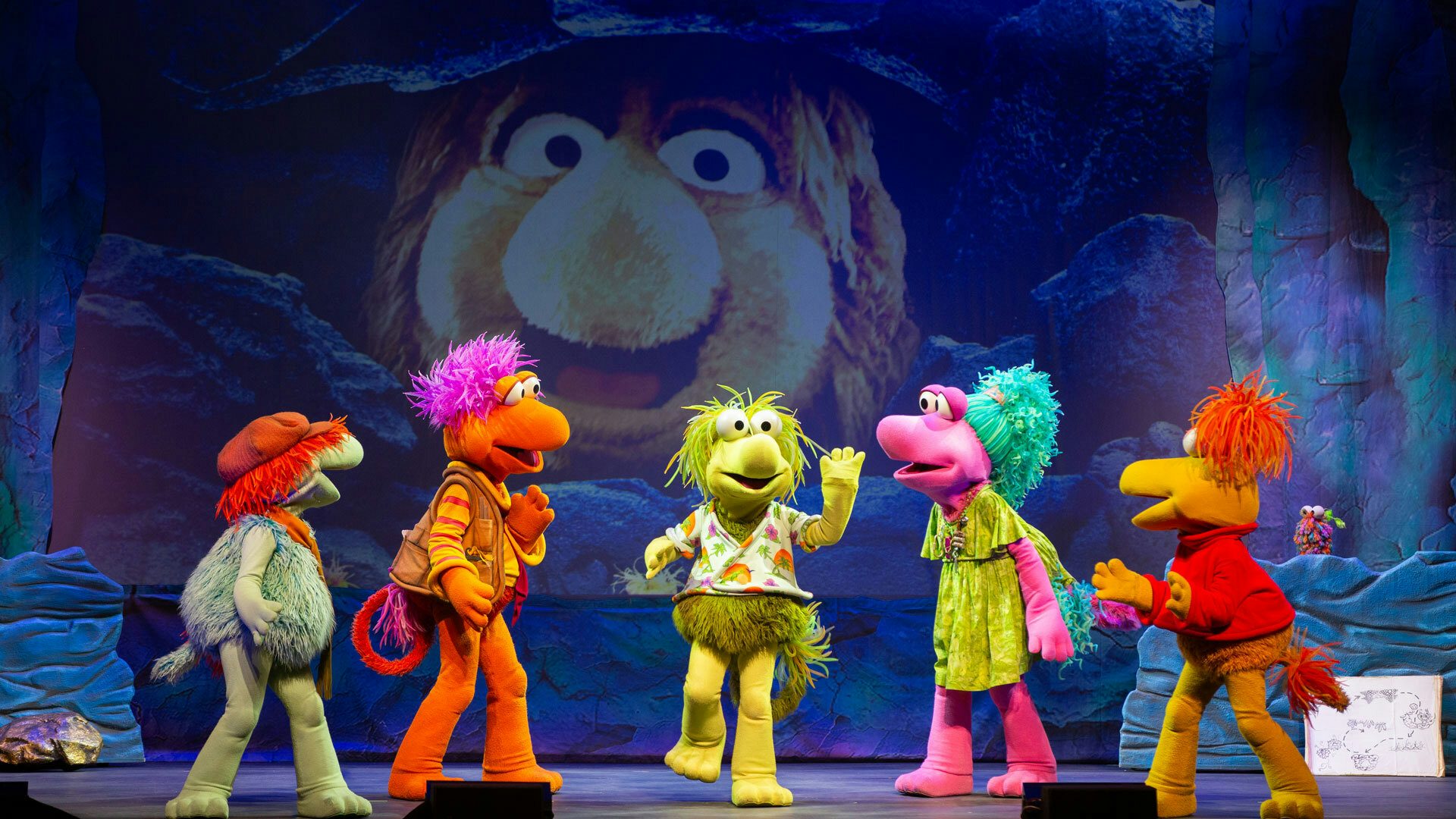 On a stage with a cave backdrop that shows a large, furry, yellow creature, five colorful Fraggles stand in a line. The teal and orange Fraggles on the left look to the right. The yellow-green fraggle in the middle raises one hand and foot while facing the audience. The pink and yellow Fraggles on the right look left.