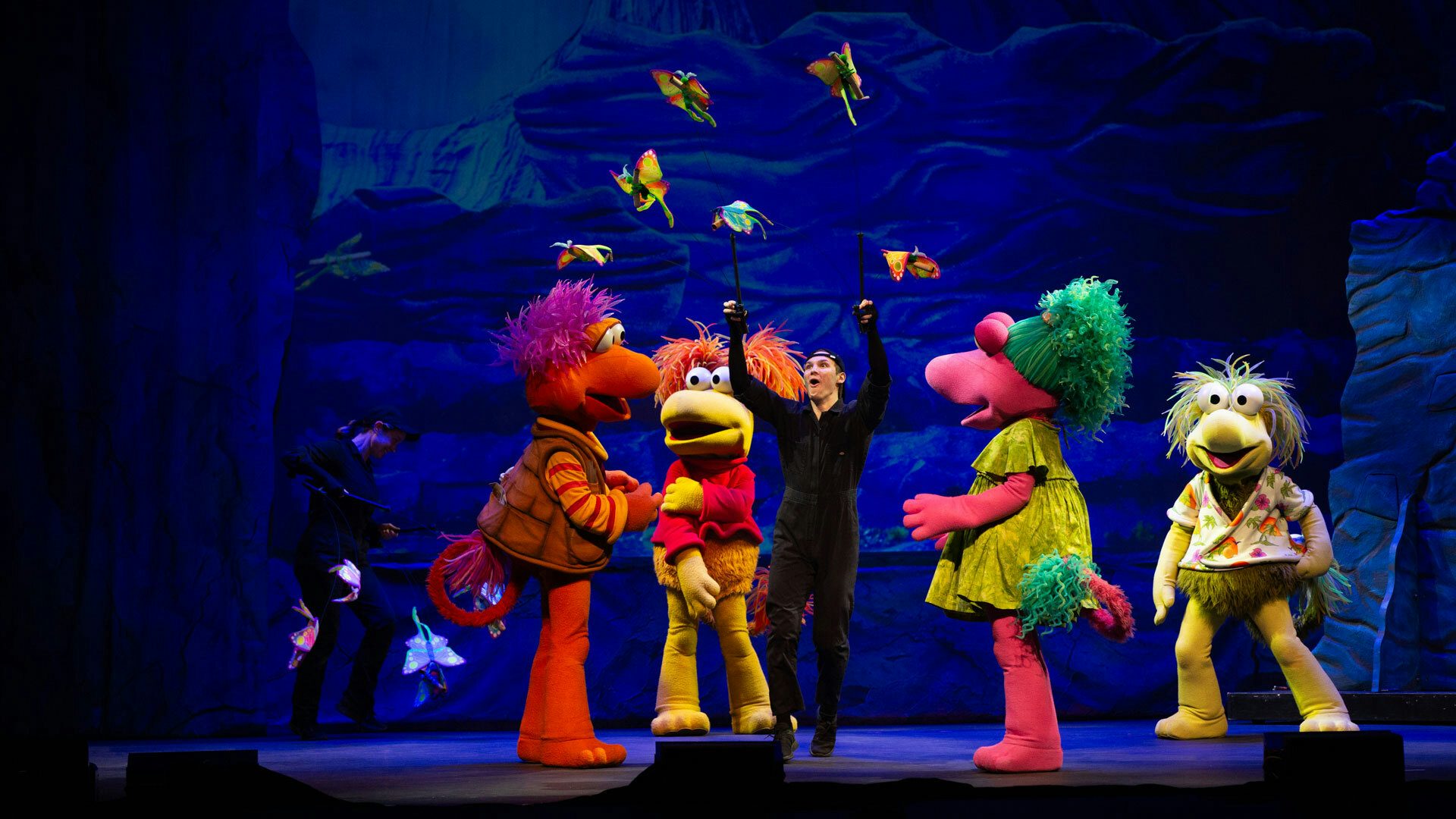 On a stage with a blue cave backdrop, two puppeteers dressed in all black perform with butterfly puppets and four colorful Fraggles stand watching the butterfly puppets around them.