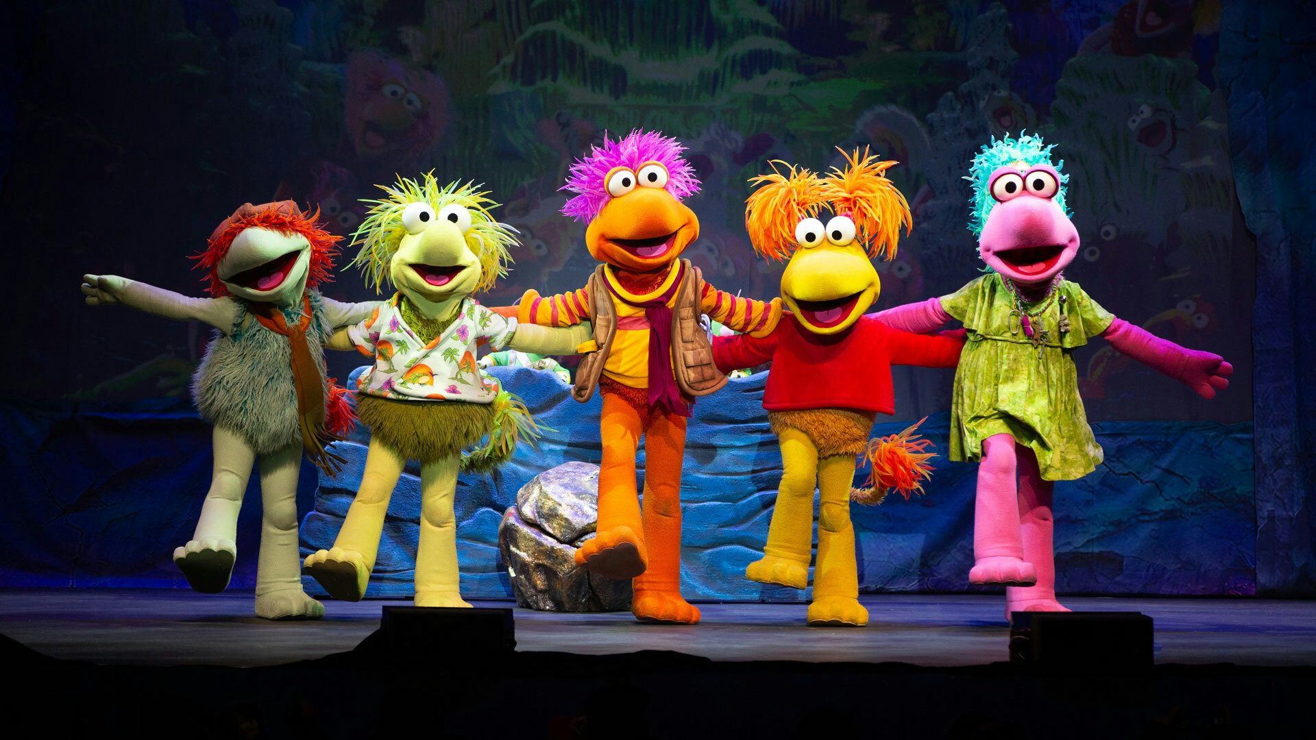 On a stage with a cave backdrop, five colorful and furry Fraggles stand in a kickline with their mouths open, arms outstretched and one foot kicked out in front of them. From left to right, the Fraggles are teal, yellow-green, orange, yellow and pink.