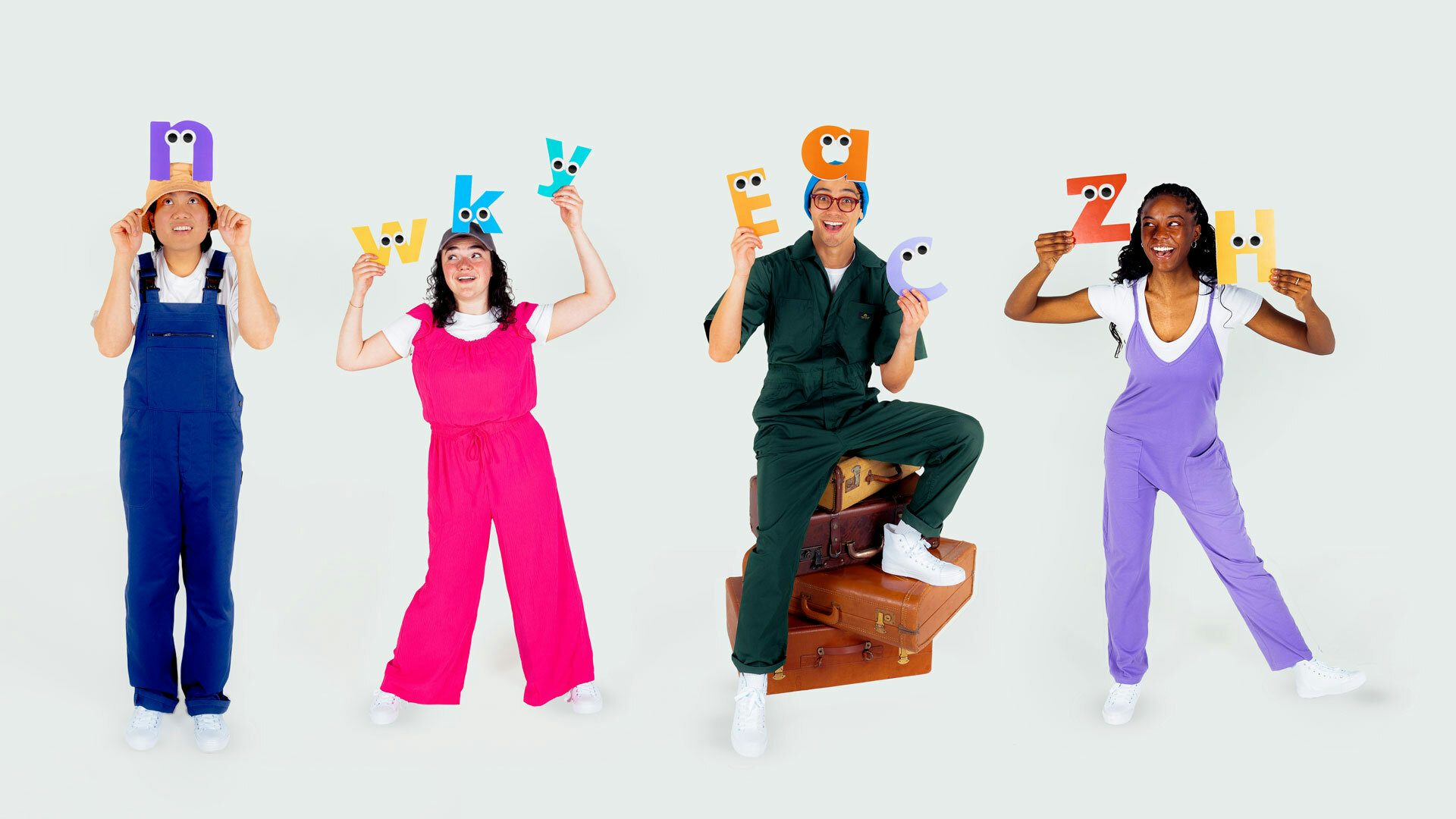 Four performers wearing colorful overalls stand smiling and holding cartoon letters with googly eyes above their heads against a white backdrop.