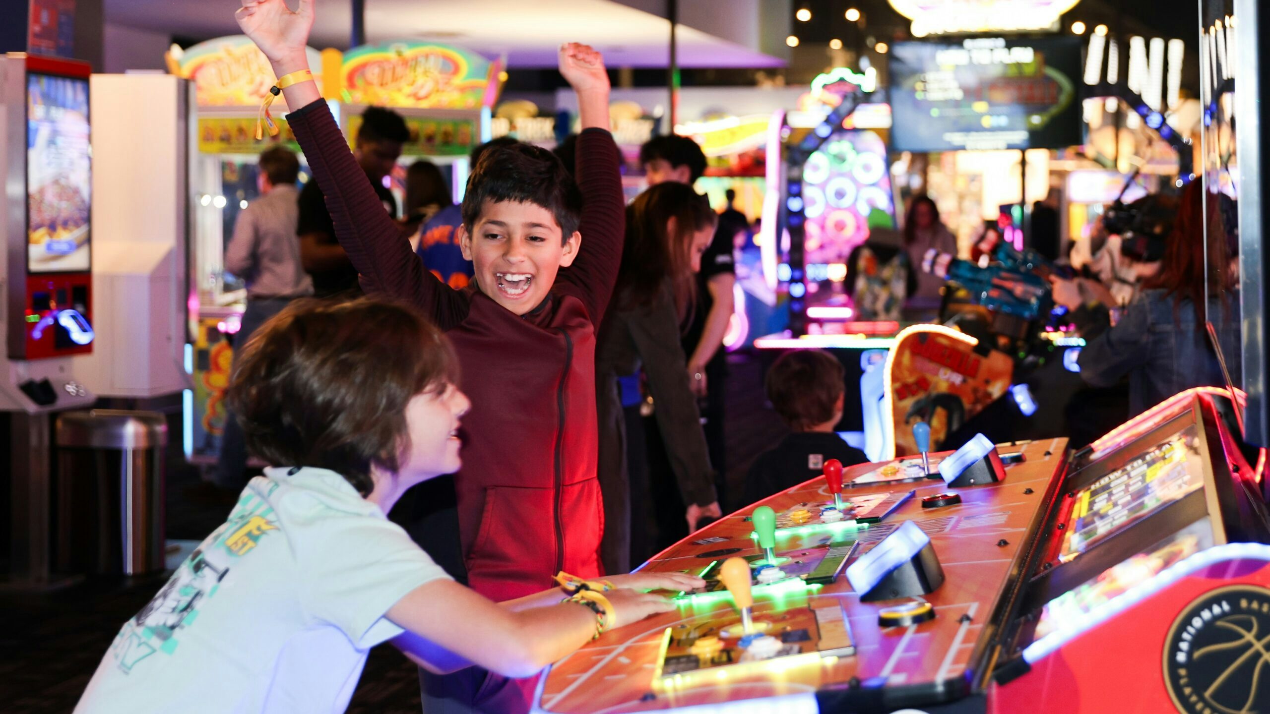 A kid wearing a red jacket throws his hands in the air and smiles at another kid to the left wearing a gray shirt playing a colorful game at Dave & Buster's.