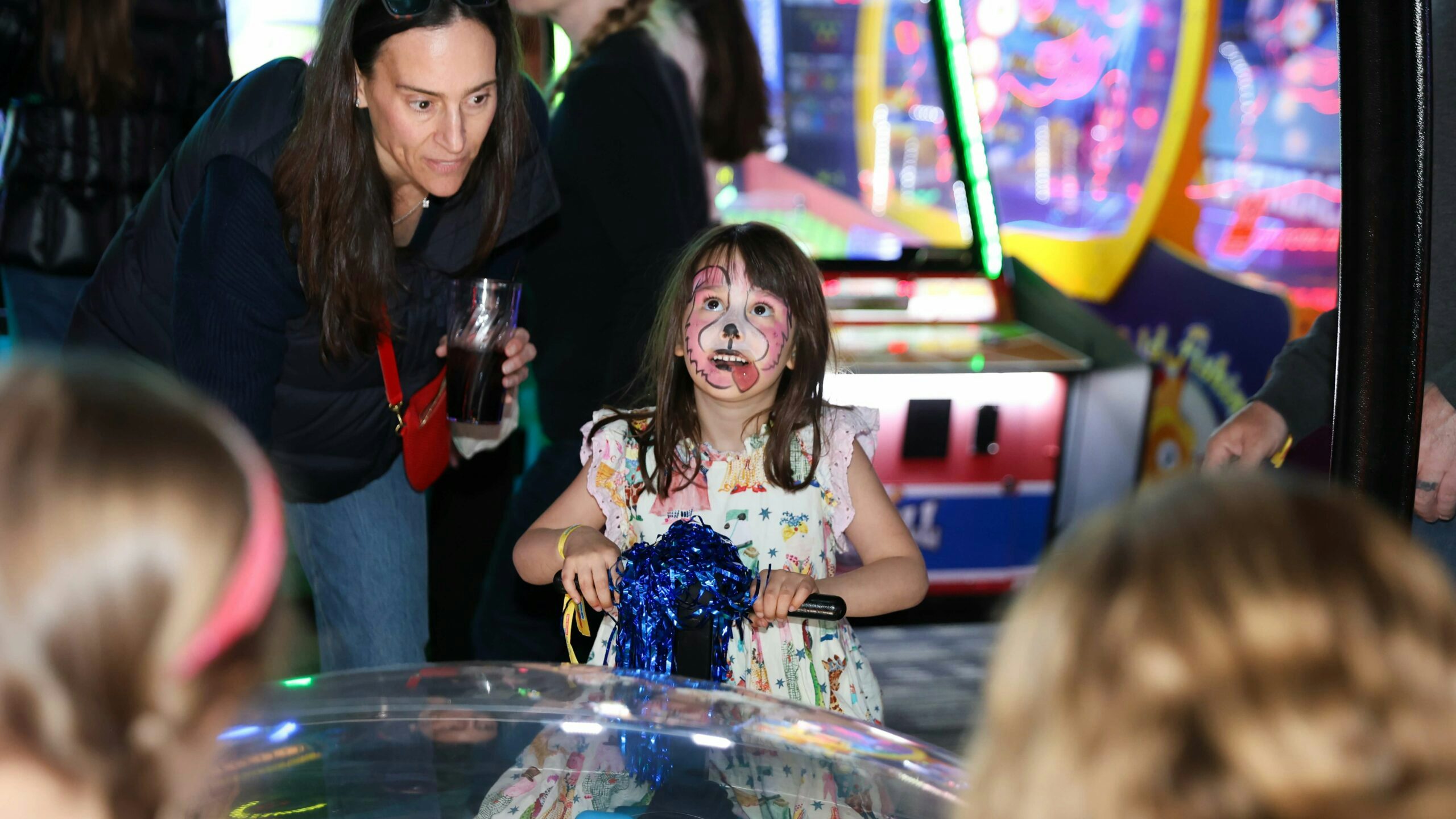 A girl with brown hair and pink puppy face paint holds a blue pom pom and looks up smiling at a Dave & Buster's game as a woman to the left wearing a dark vest and holding a glass looks on.