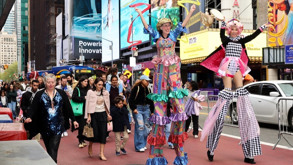 Two stilt walkers in colorful clothes lead a group of kids and adults down 42nd Street.