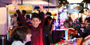A boy in a red jacket having his arms raised in excitement at an arcade.
