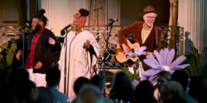 Three performers singing in front of a crowd. One performer is also playing the guitar. 