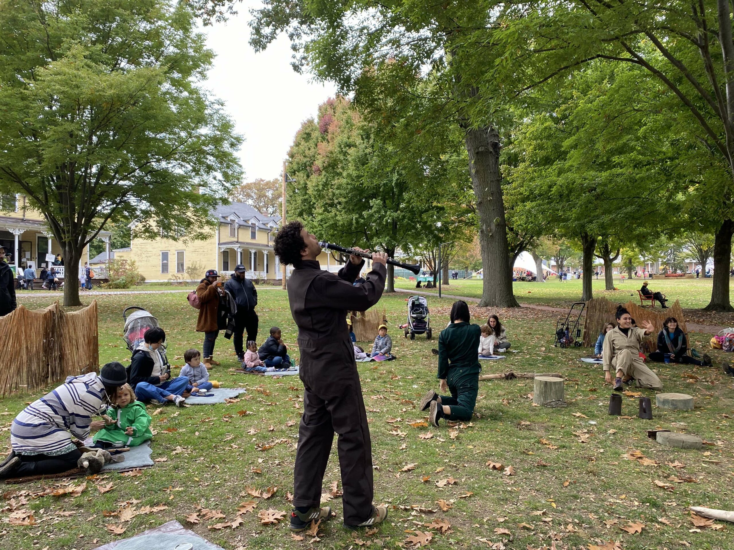 A person plays a clarinet outdoors in a park surrounded by families sitting on blankets. Trees and a building are in the background. The atmosphere is relaxed and communal.