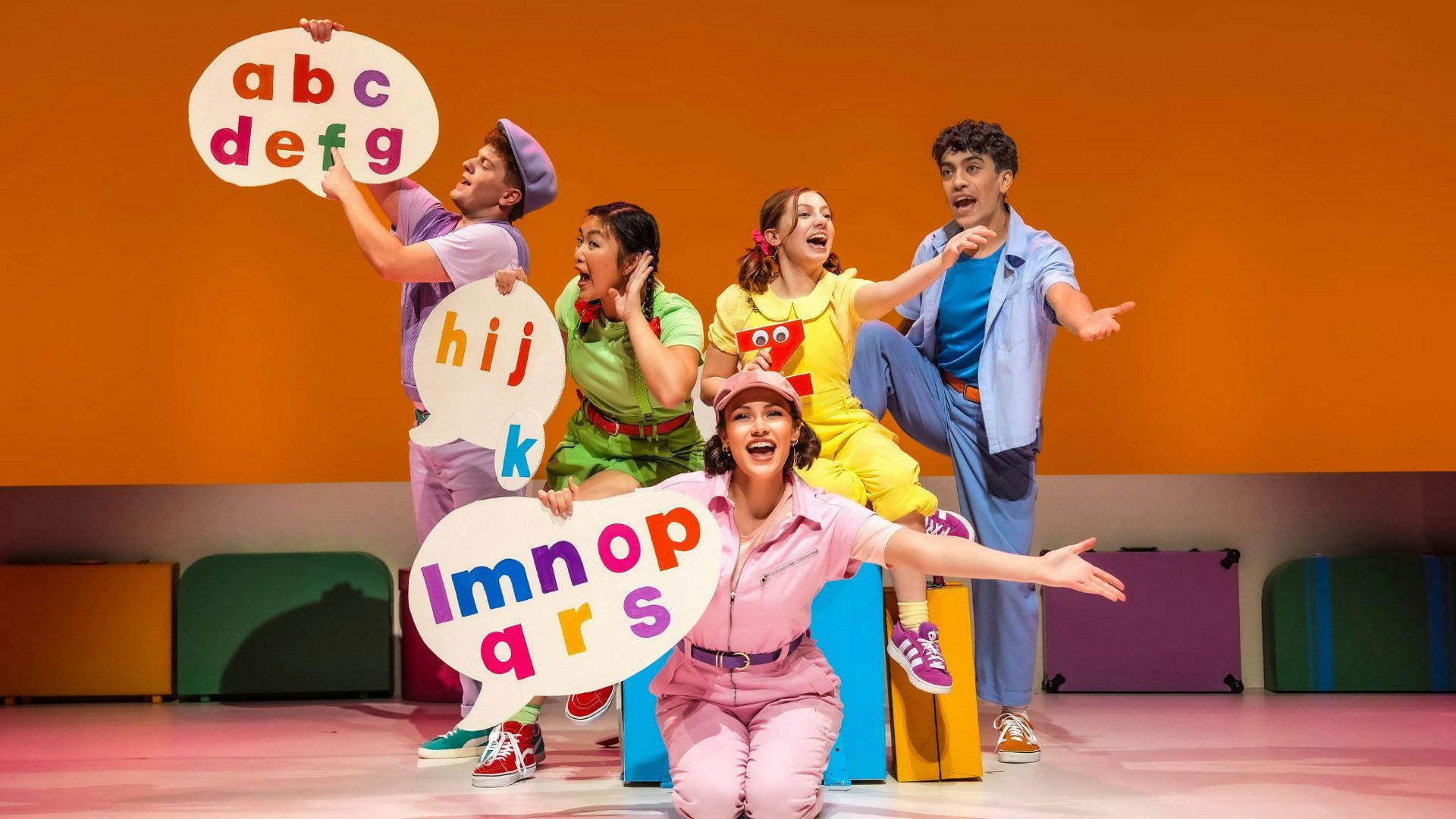 Five performers in colorful clothing hold speech bubbles with letters of the alphabet on them and gesture for the audience to sing along.