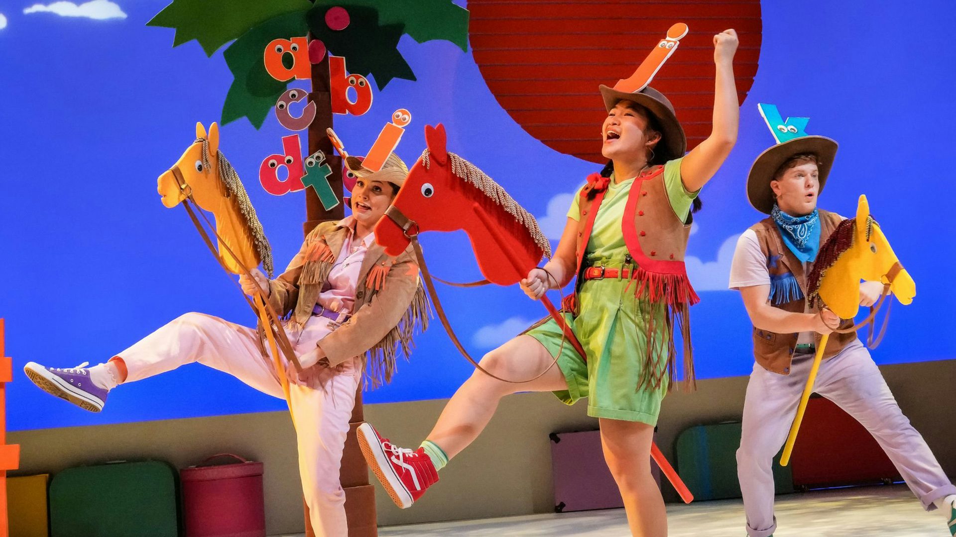 Three performers in fringe clothing ride hobby horses. They each wear cowboy hats with a different lowercase letter on them.