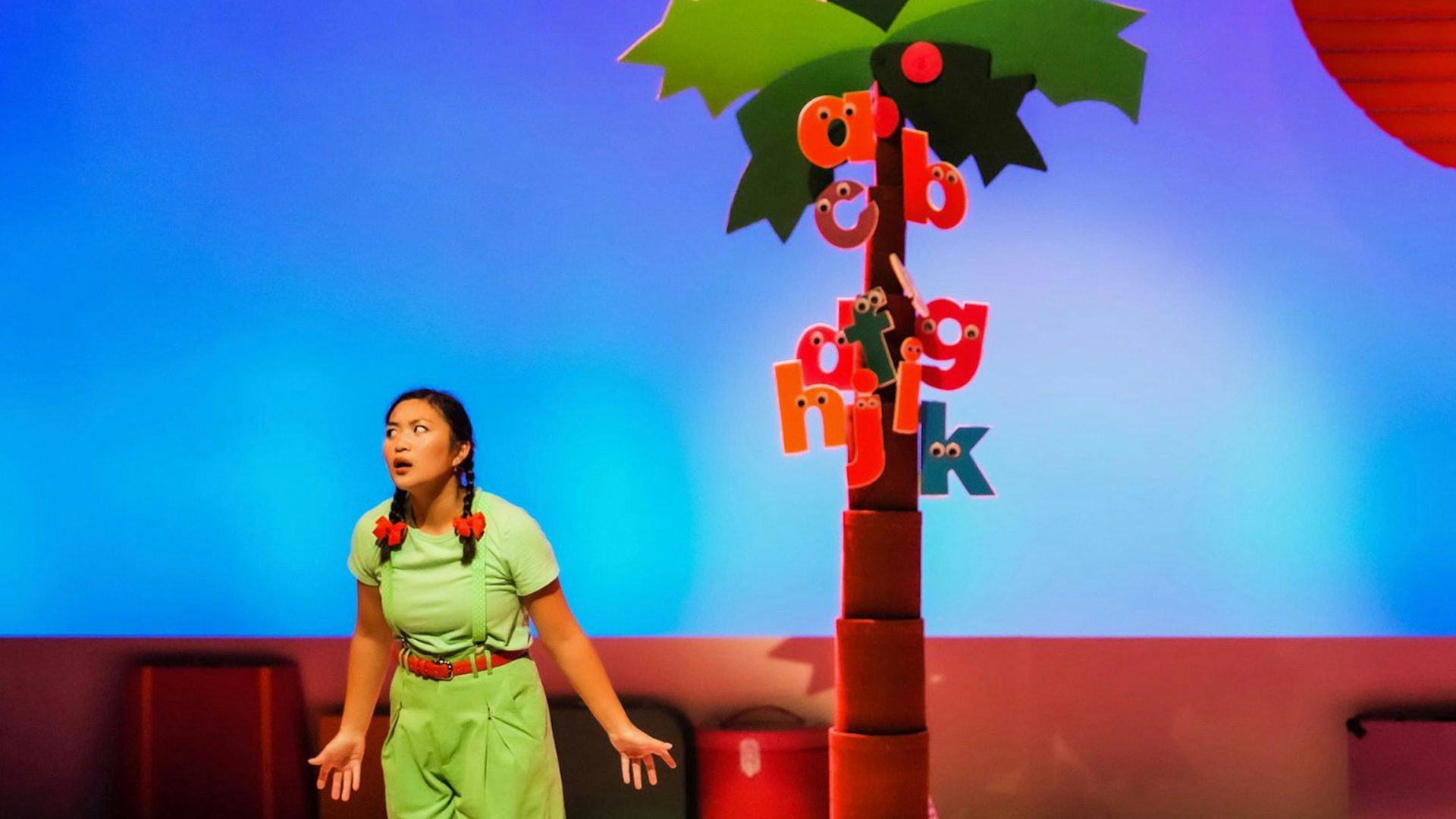 A performer wearing green makes a confused expression. Behind the performer, lowercase letters with googly eyes cling to the trunk of a coconut tree.