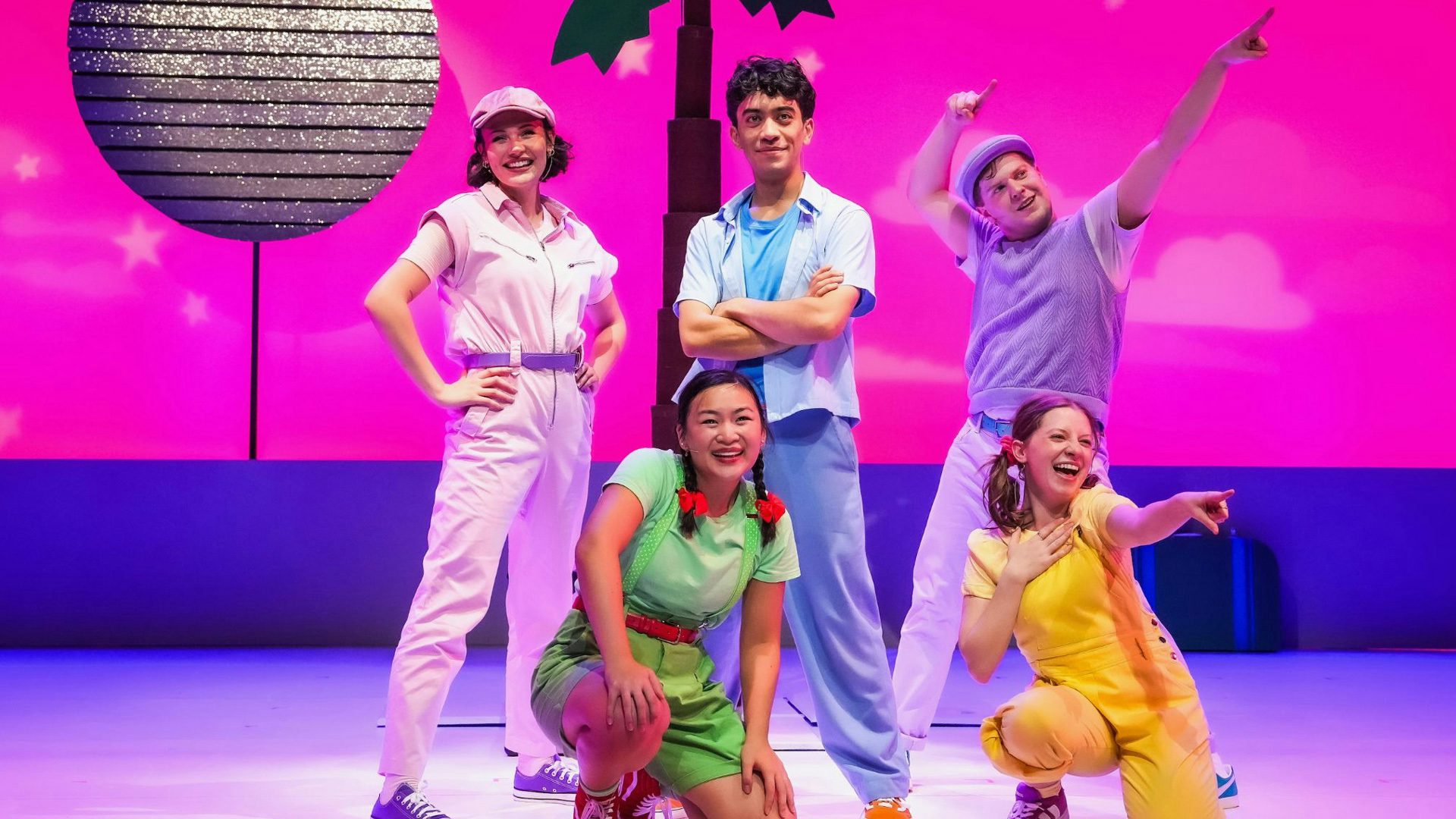Five performers in colorful clothing smile and strike a pose on stage in front of a coconut tree and sparkly moon on a pink sky backdrop.