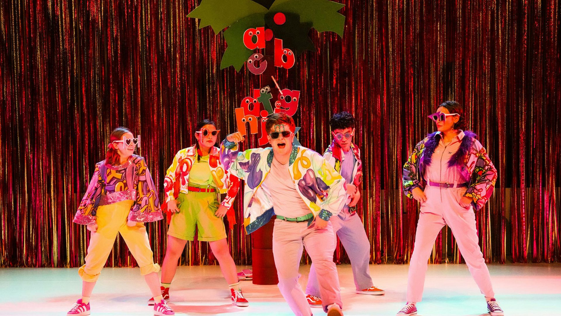 Five performers in colorful jackets and sunglasses dance in front of a coconut tree and fringe backdrop. Lowercase letters with googly eyes cling to the tree's trunk.