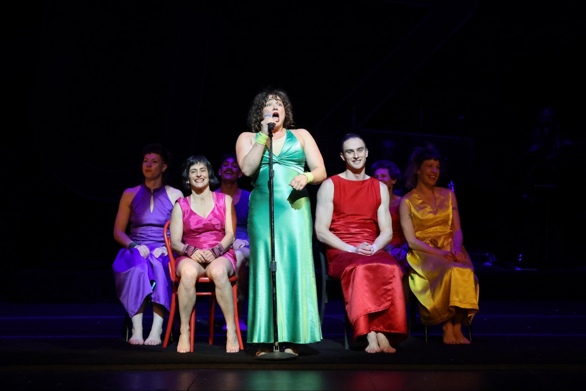 A performer in a long green dress speaks into a microphone while other performers sit in chairs, smiling.