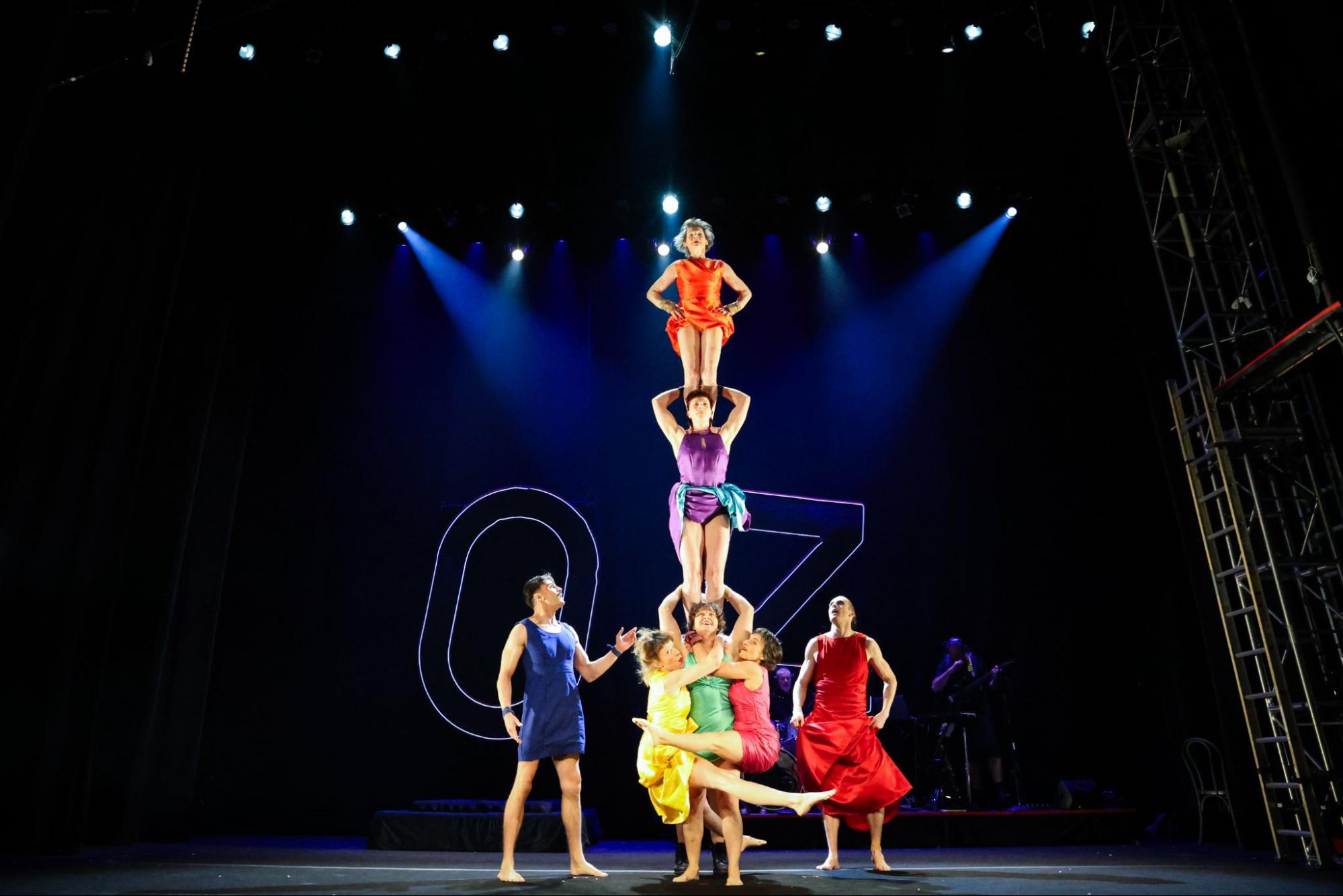 Two people in colorful dresses stand on top of a third person who has two other people hanging off the ground by clinging on to her.