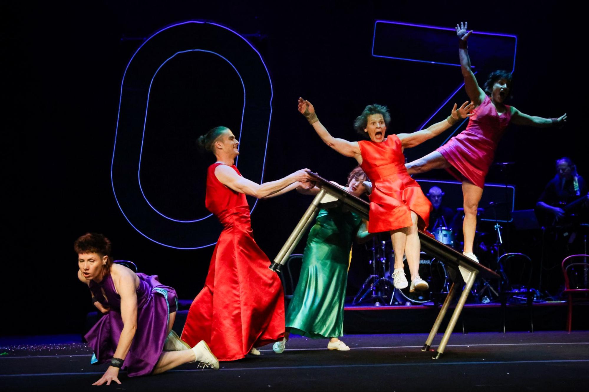 Several performers are surprised as they slide off a table lifted but another performer.