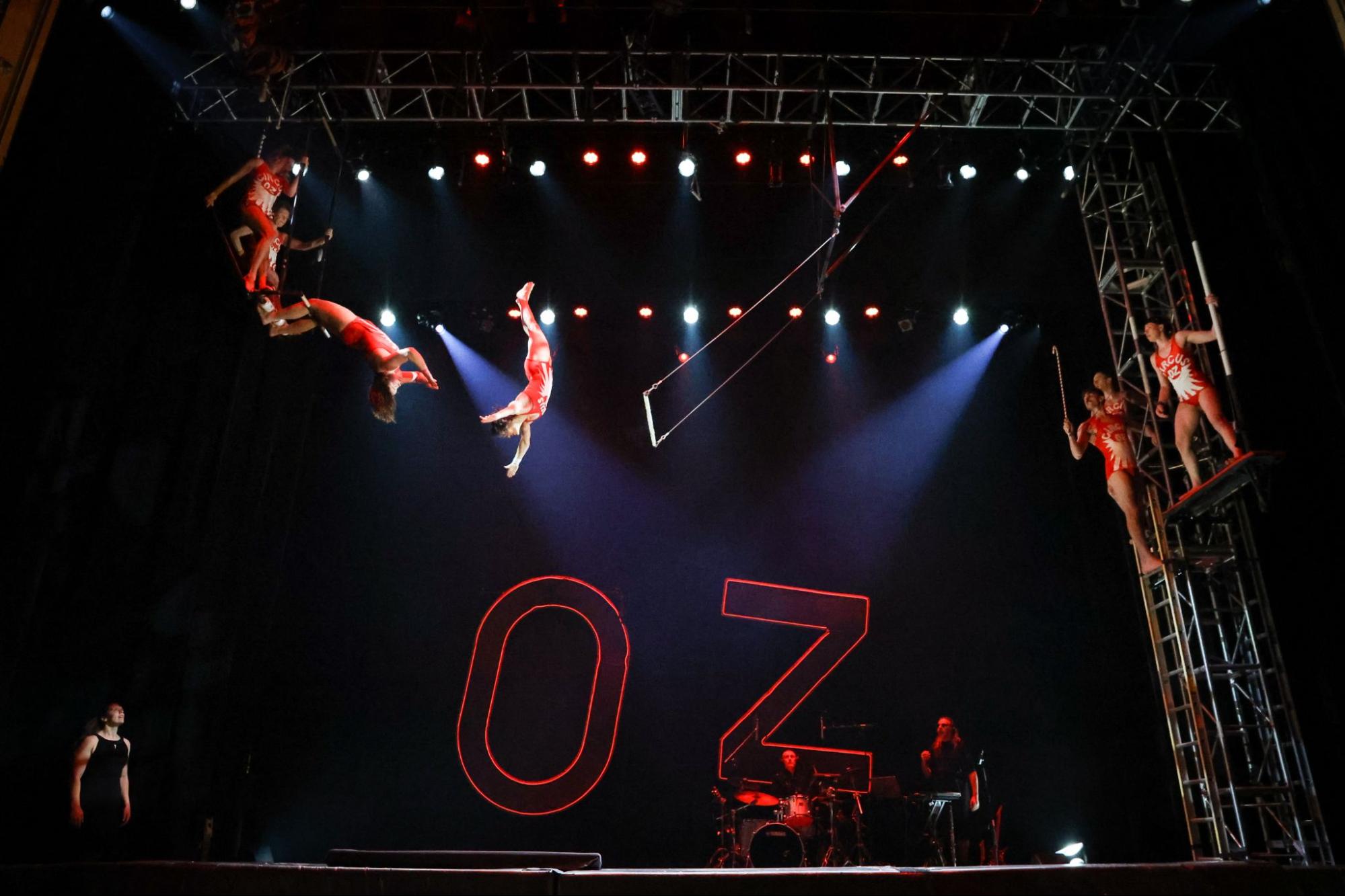 Performers watch to the right and left while a trapeze artist is in the middle of a flip off of a trapeze swing towards a performer bending backwards off of a bar to catch the trapeze artist.